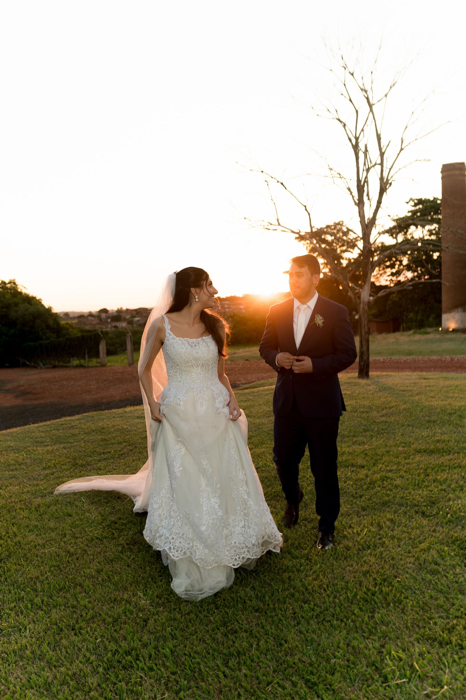 Bride and groom walking on green lawn at golden sunset, bride in lace wedding dress with veil, groom in dark suit.