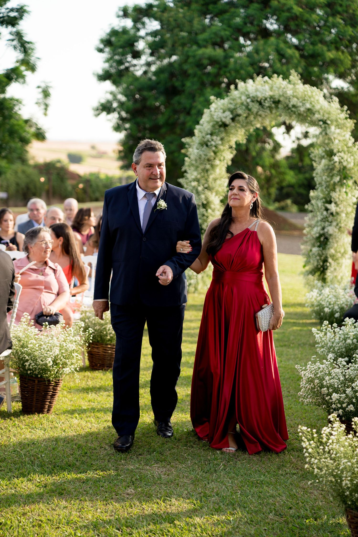 Man in navy suit and woman in red gown walk down outdoor wedding aisle lined with white floral arrangements.