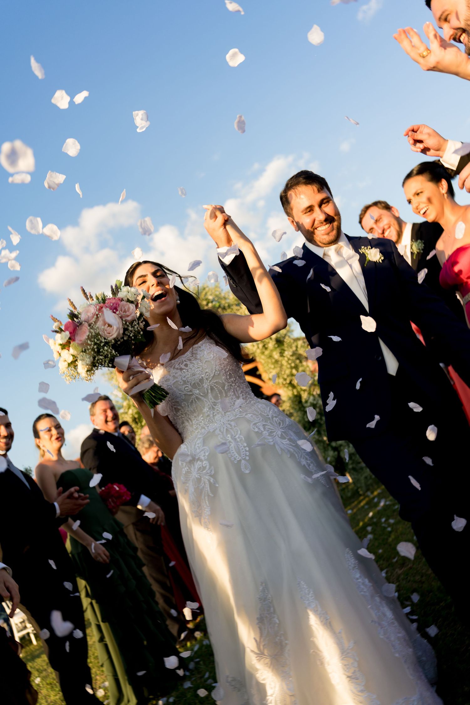 Joyful bride and groom walk down aisle as guests toss white rose petals at outdoor wedding ceremony.