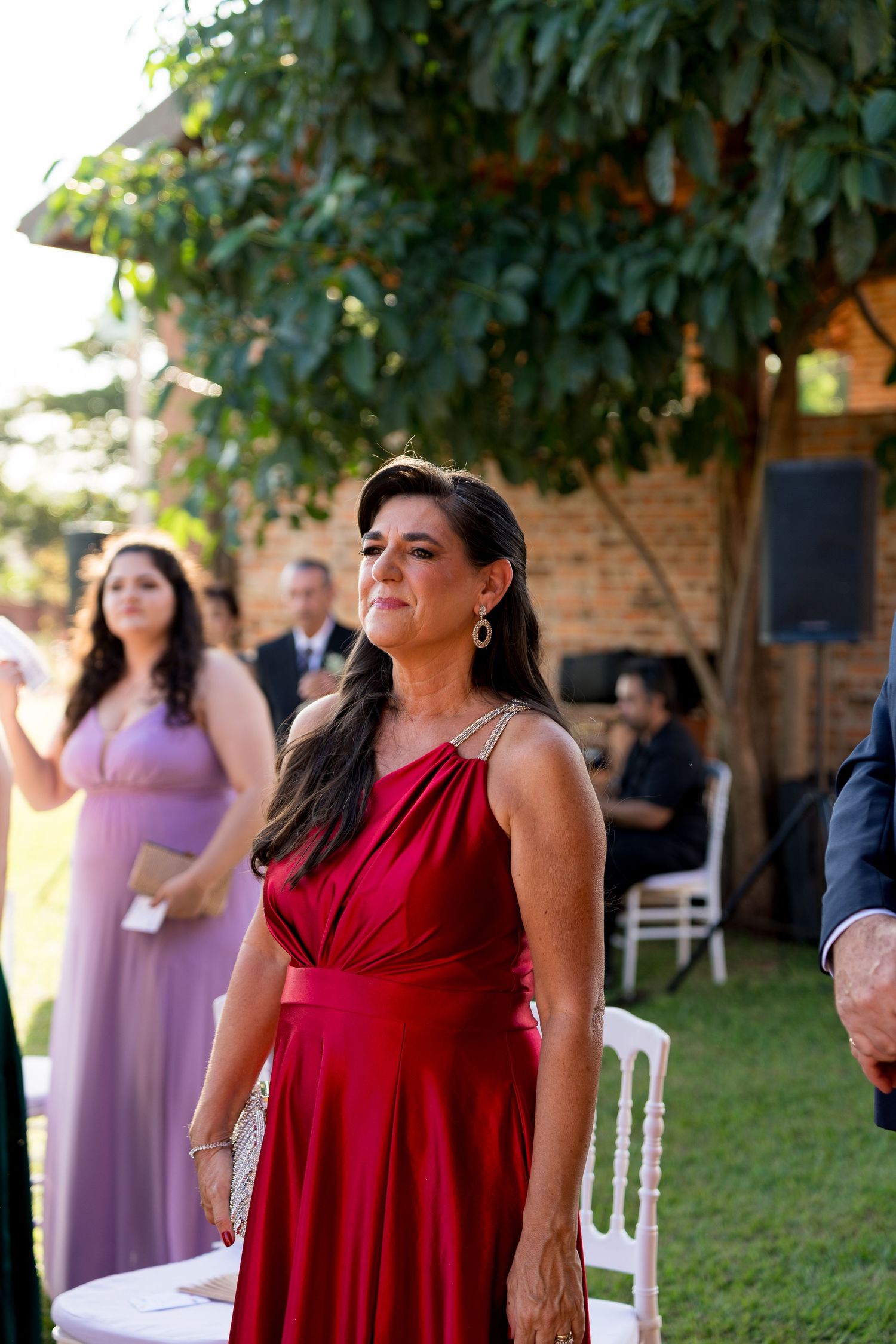 Woman in red gown smiling at outdoor wedding ceremony with guests in background.