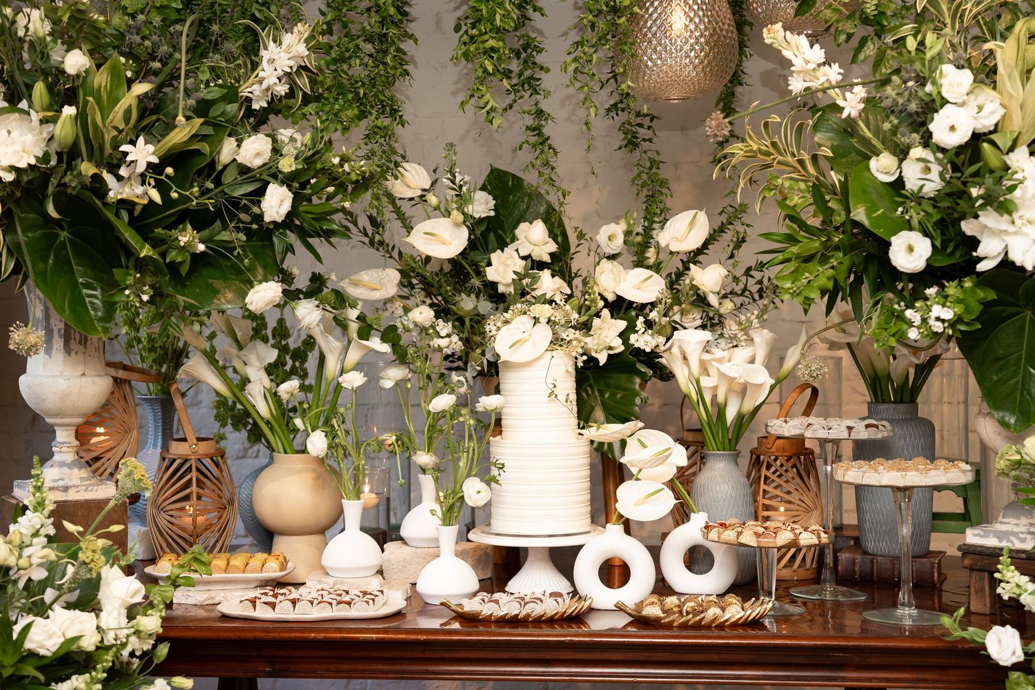 Elegant wedding dessert table with white floral arrangements, tiered cake, and decorative sweets on a wooden table.
