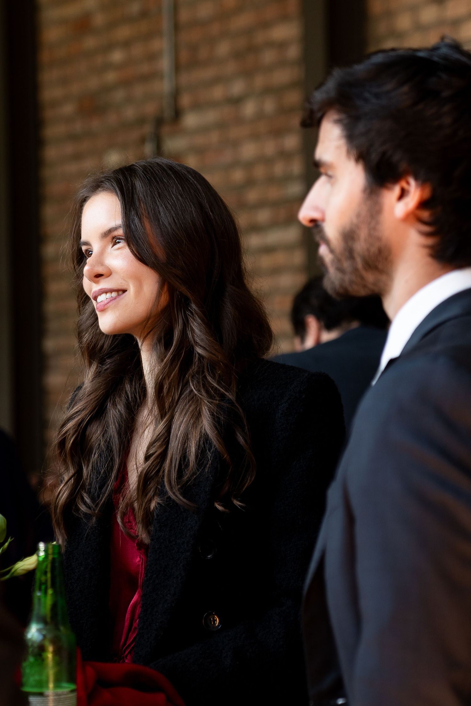 A smiling brunette woman in a black coat stands beside a bearded man in a suit near a brick wall.