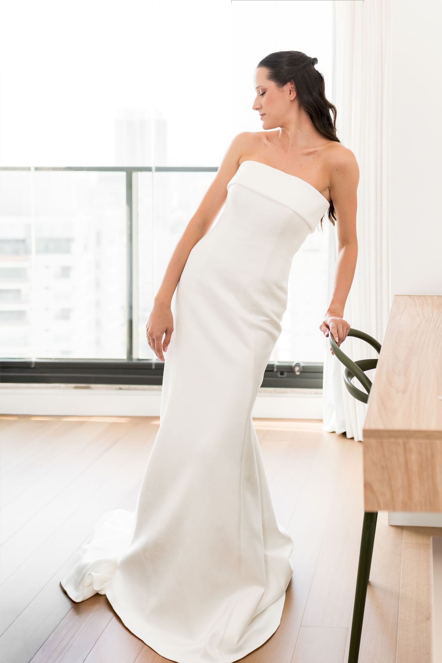 Bride wearing a sleek strapless white mermaid wedding gown with a train, standing by large windows on hardwood floors.