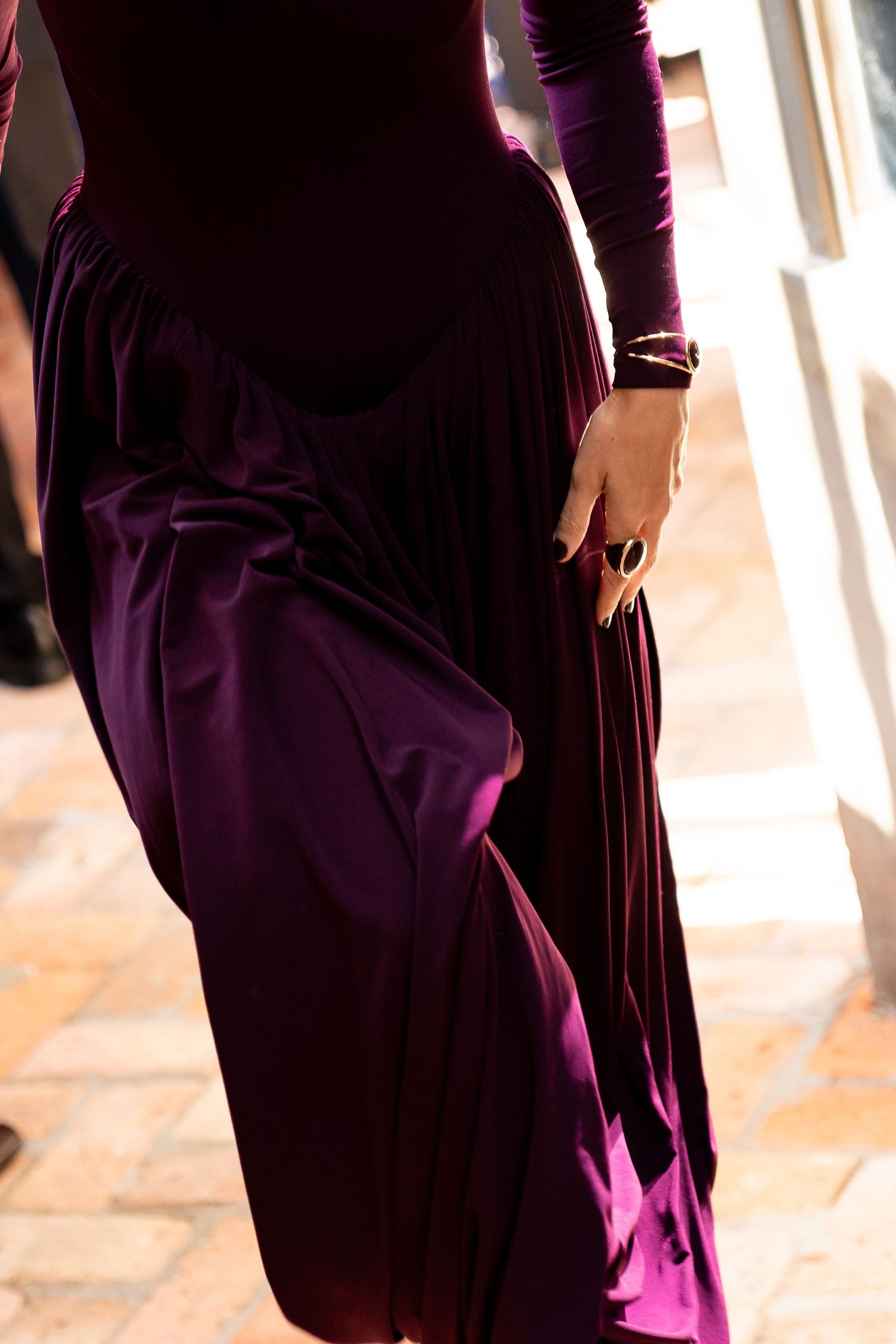 Elegant woman in a deep purple velvet gown with gold bracelet and ring, holding skirt on tiled floor.