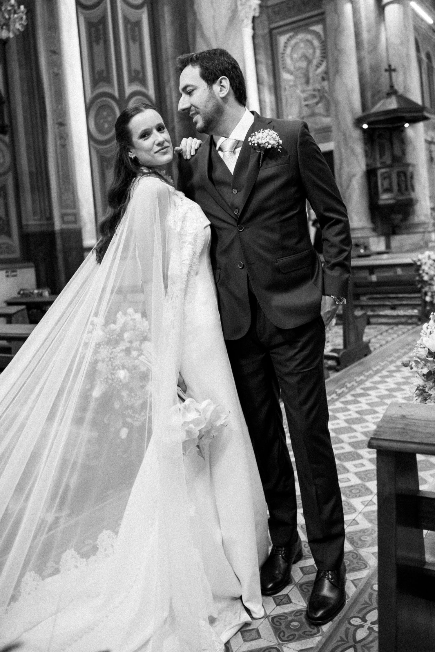 Black and white wedding photo of bride in lace gown with veil and groom in dark suit embracing in ornate church.
