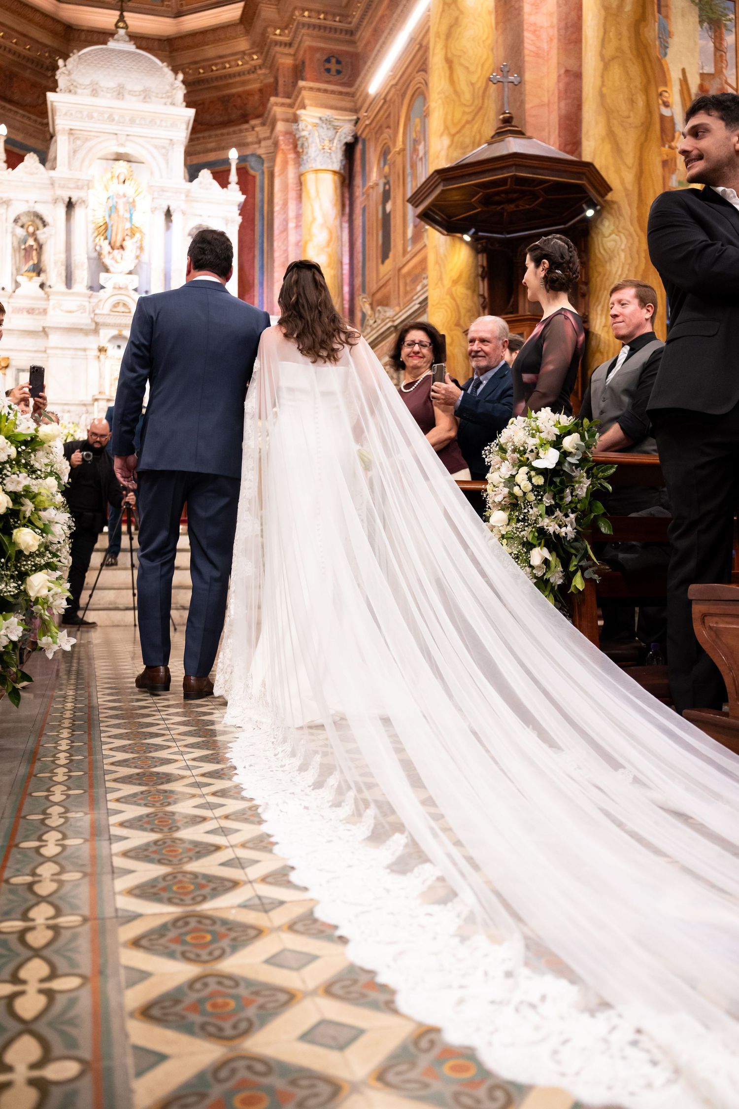 Bride in white gown with long lace veil walks down church aisle with groom in blue suit at wedding ceremony.