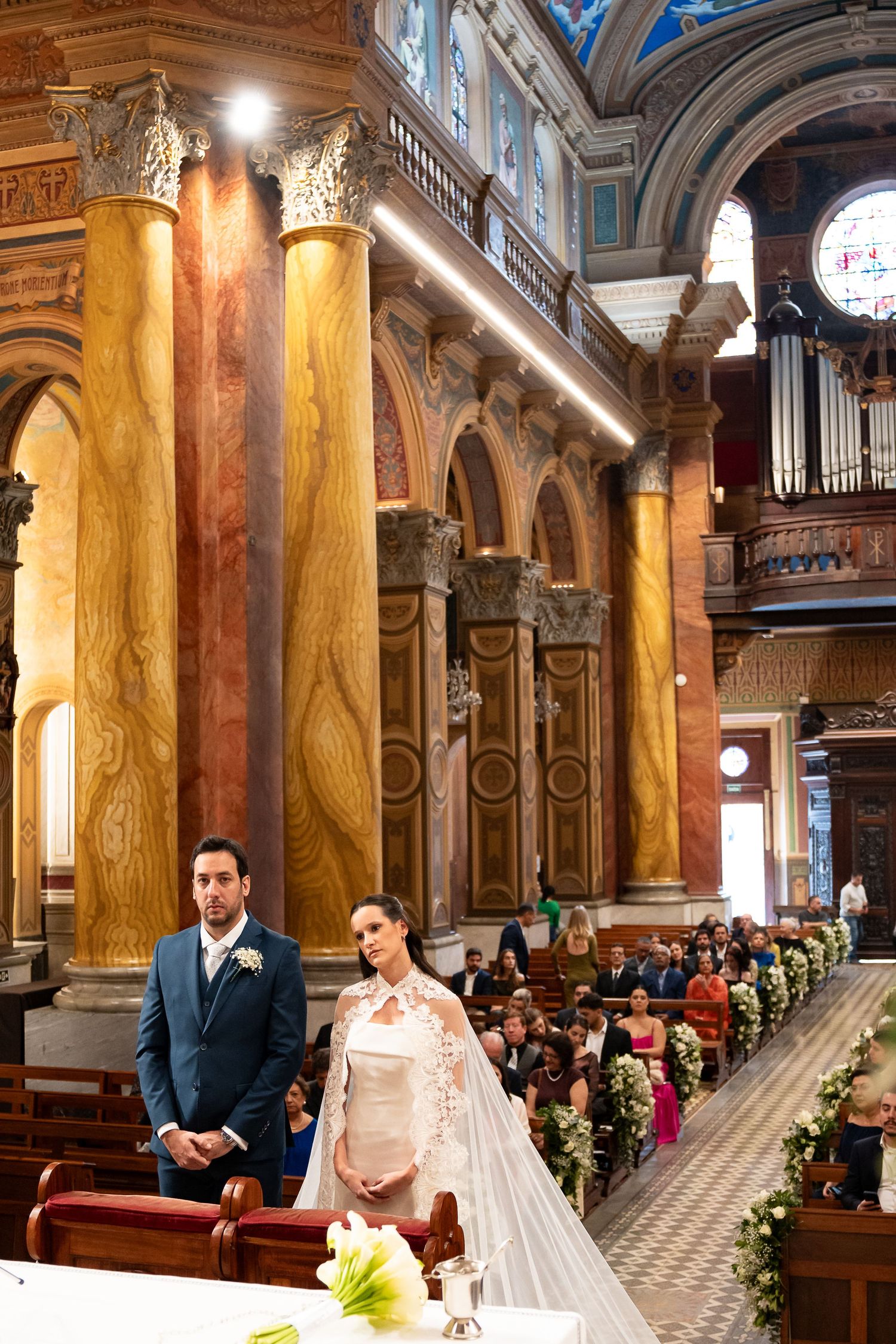 Bride and groom stand at the altar of an ornate church with grand columns during their wedding ceremony.