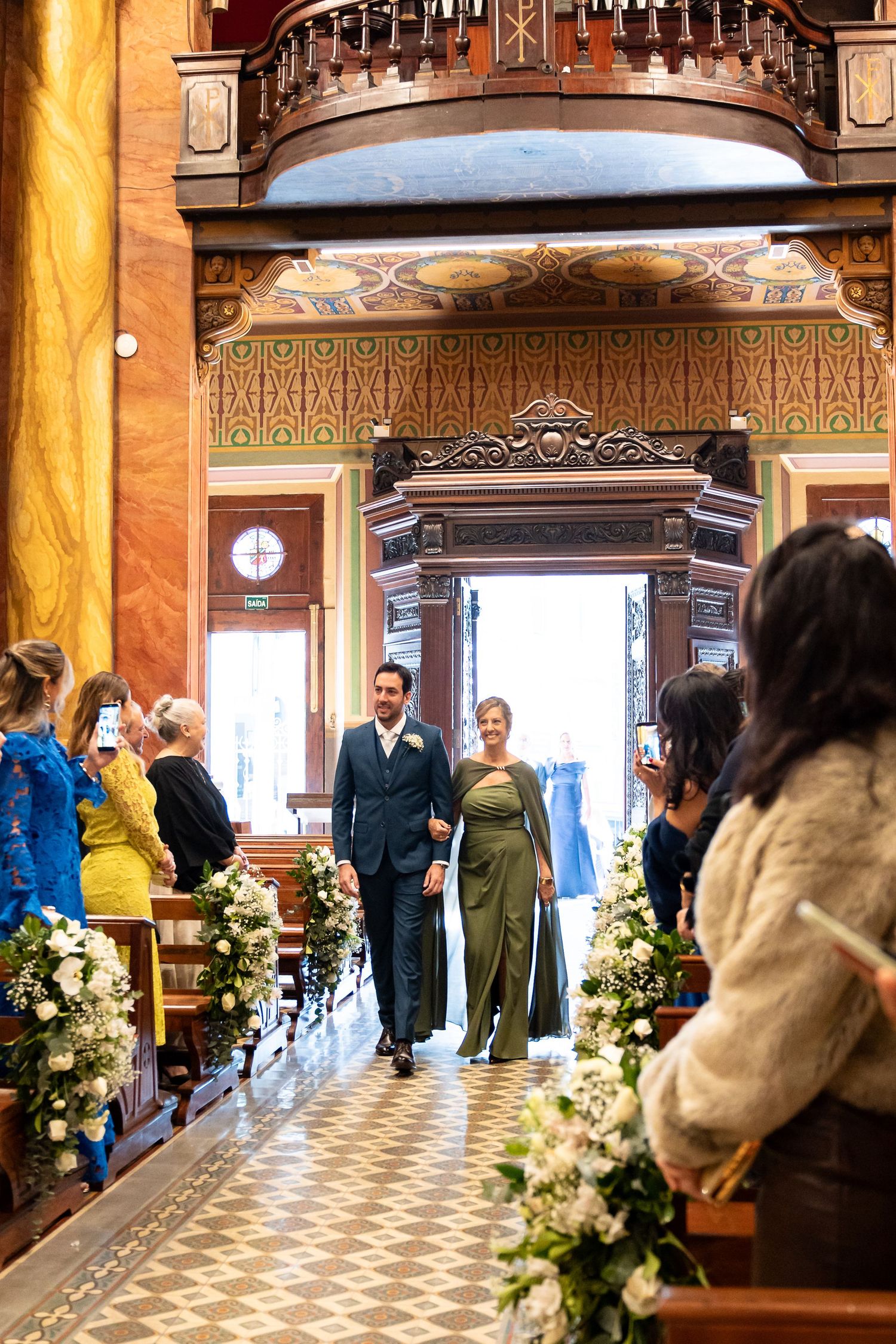 Couple walks down ornate church aisle lined with white floral arrangements during elegant wedding ceremony.