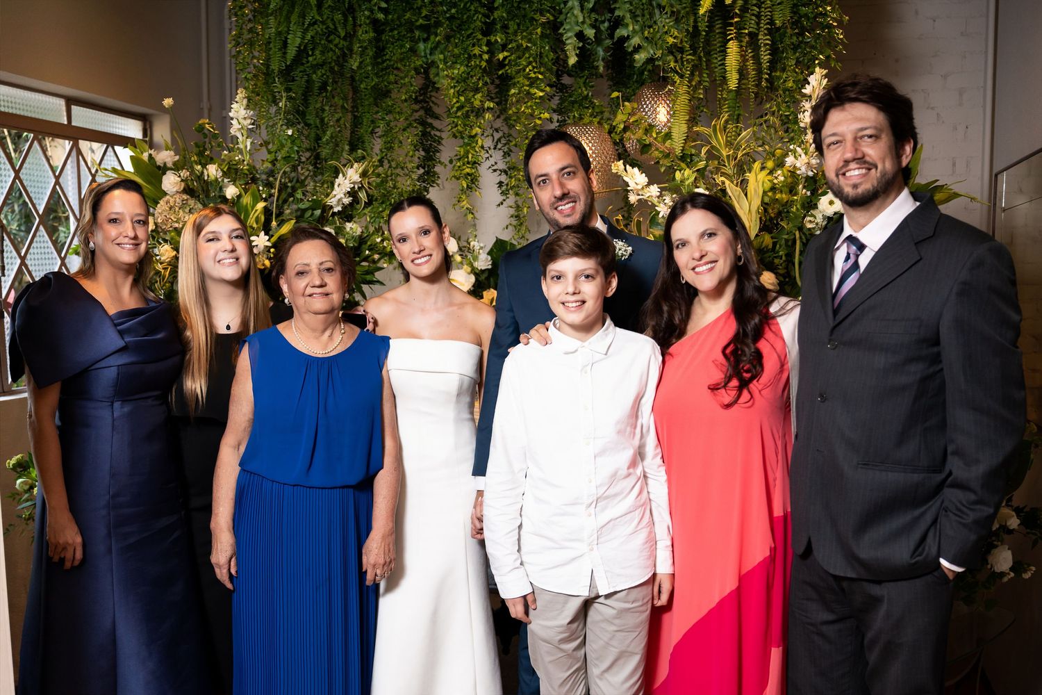 Wedding group photo with bride in white gown, groom, and family members posing before floral greenery backdrop.