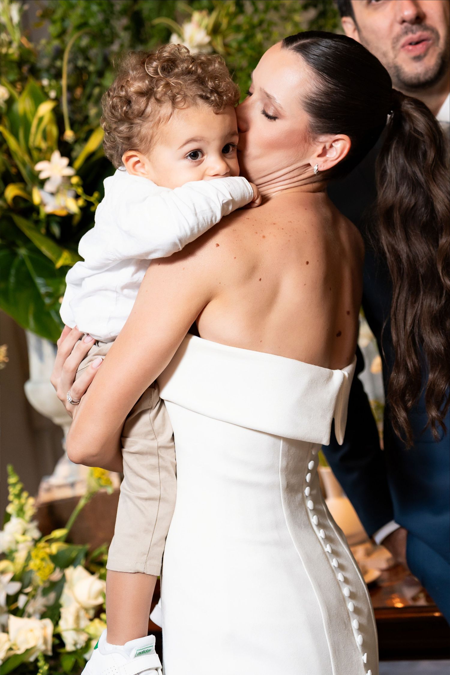Bride in white strapless gown tenderly kisses a toddler boy she holds at a wedding ceremony.