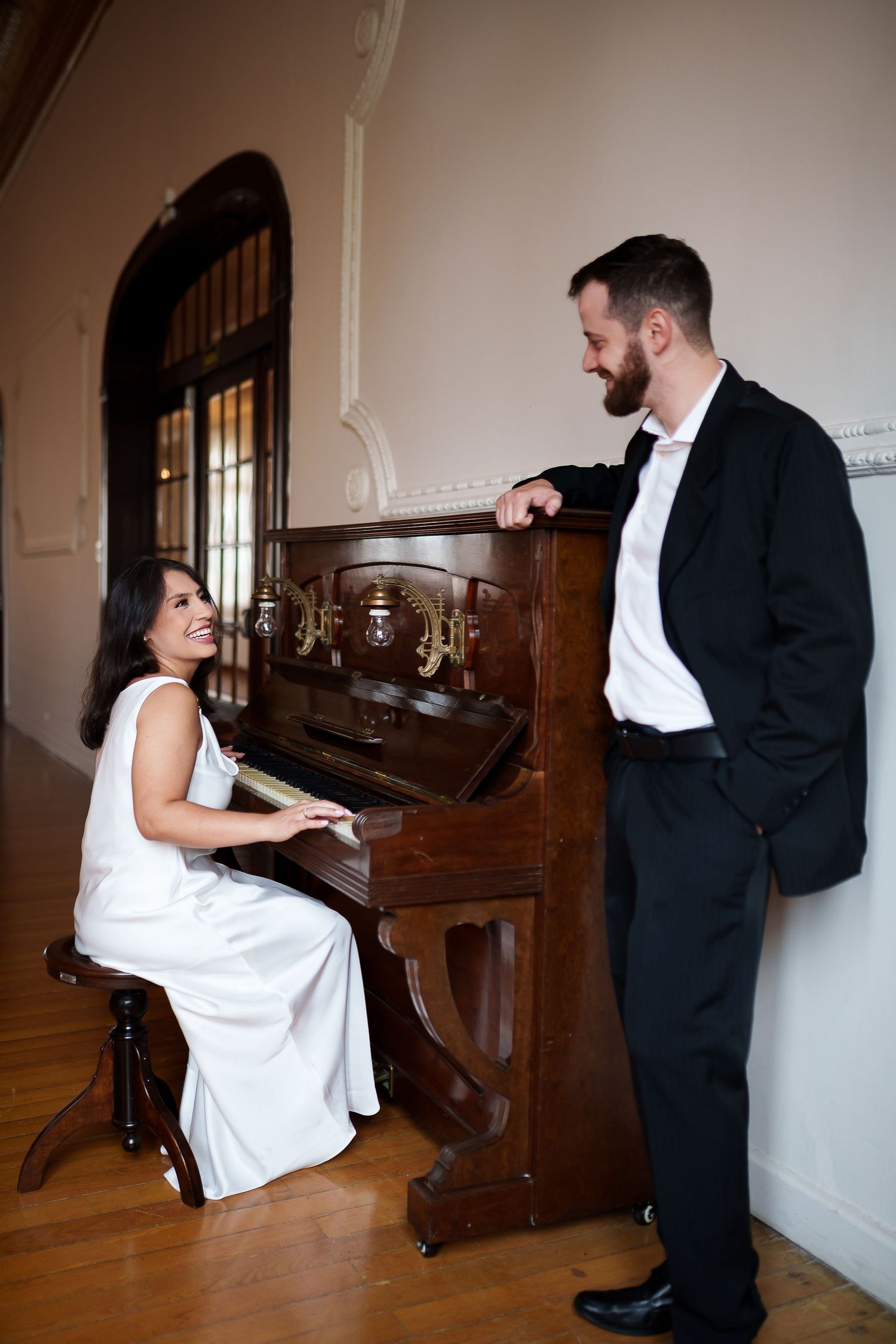 Elegant couple in formal wear, woman in white dress playing upright piano while man in tuxedo leans against it smiling.
