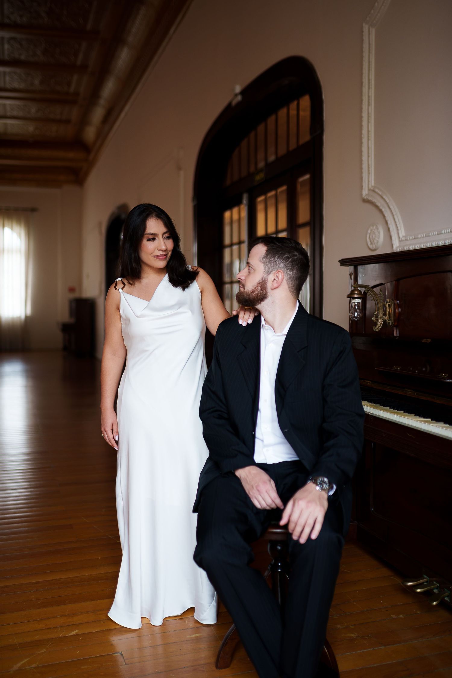 Elegant couple in wedding attire posing near a grand piano in a ornate ballroom, bride in white gown, groom in black suit.
