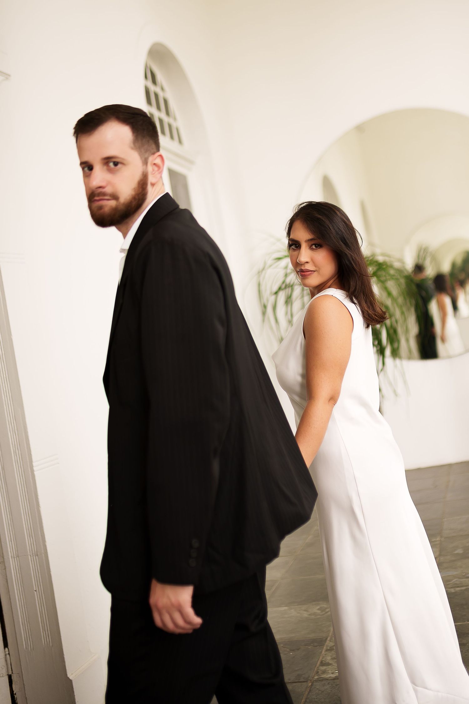 Elegant couple in formal attire, man in black suit and woman in white dress, posing in a white archway venue.