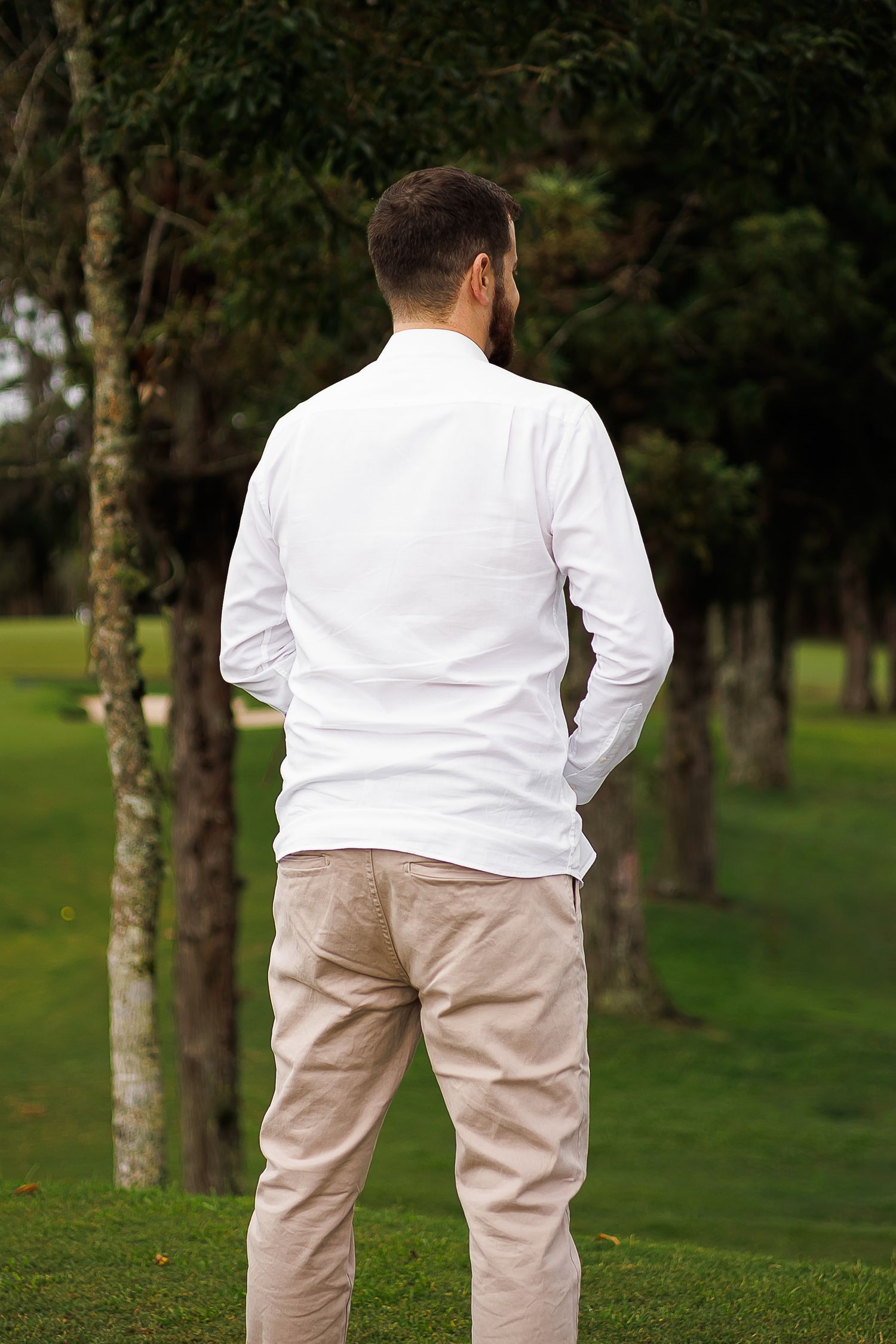 Man in white long-sleeve shirt and beige pants standing outdoors on green grass near trees, viewed from behind.