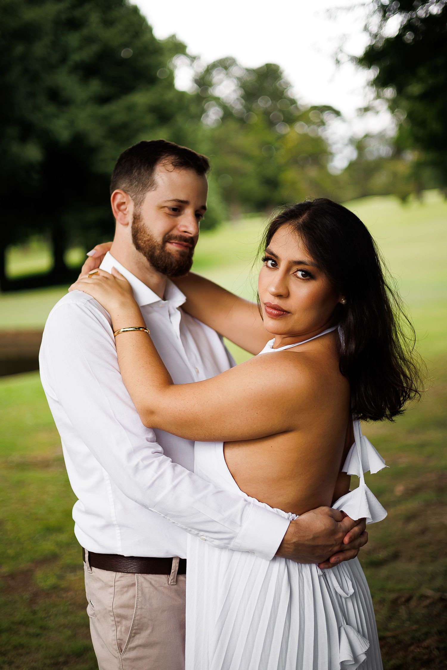 A couple in white outfits embrace in a lush green park, the woman gazing at the camera while the man holds her close.