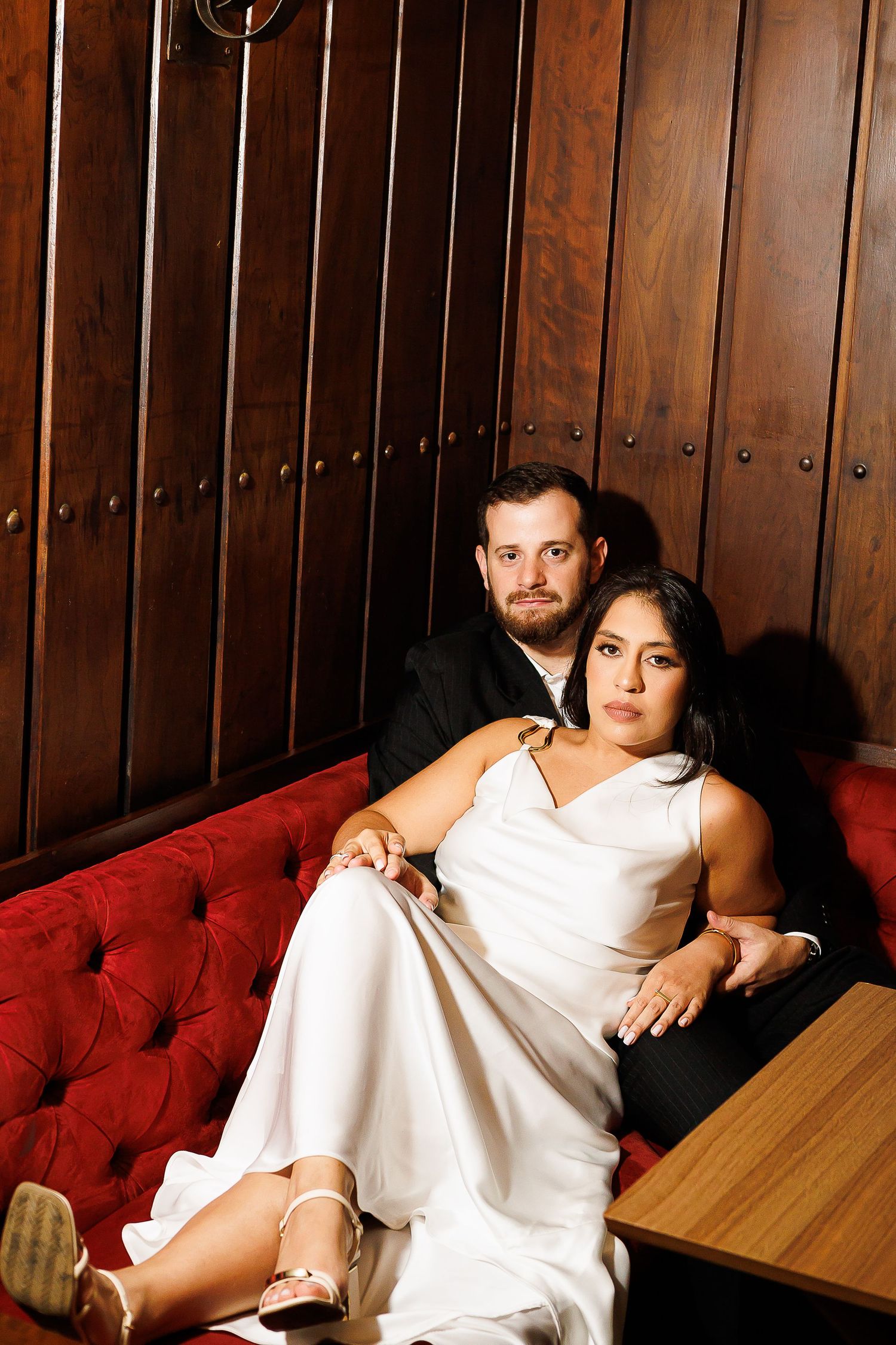 Bride and groom sitting together on a red tufted sofa in front of wood-paneled walls at their wedding reception.