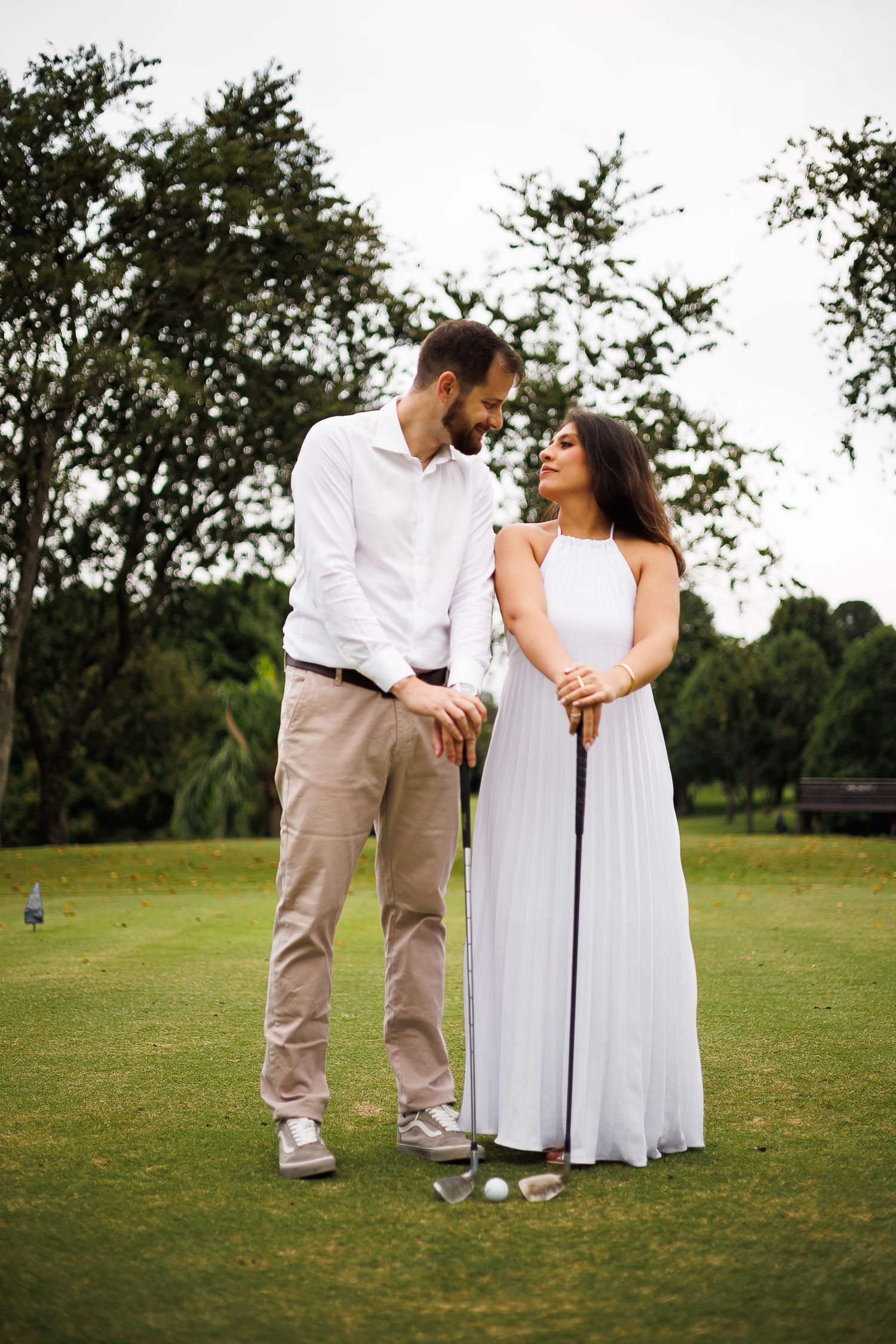 Couple in white outfits sharing a romantic moment on a golf course, woman in elegant dress holding a golf club.