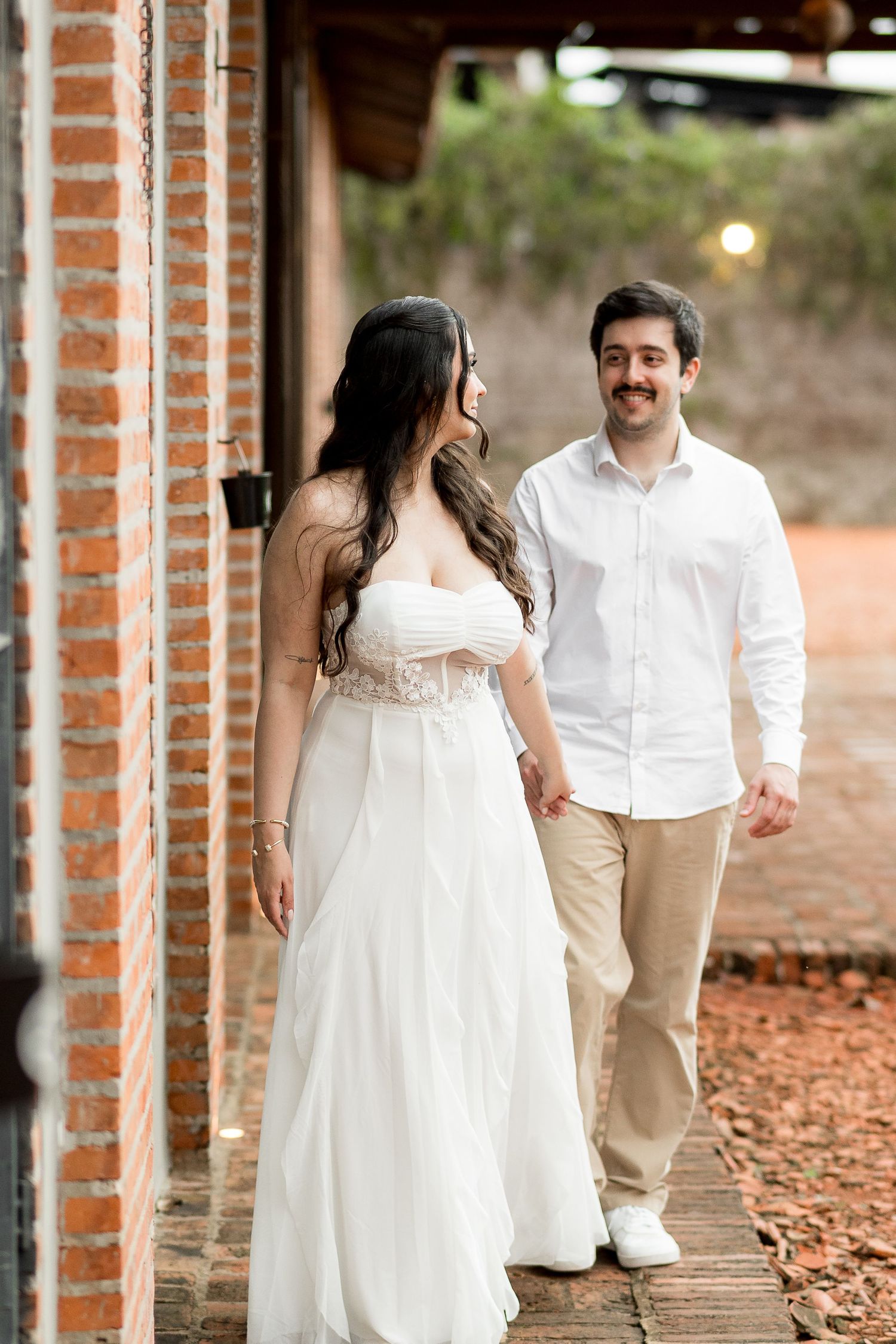 Bride in white strapless gown and groom in white shirt walk hand-in-hand along a brick building pathway.