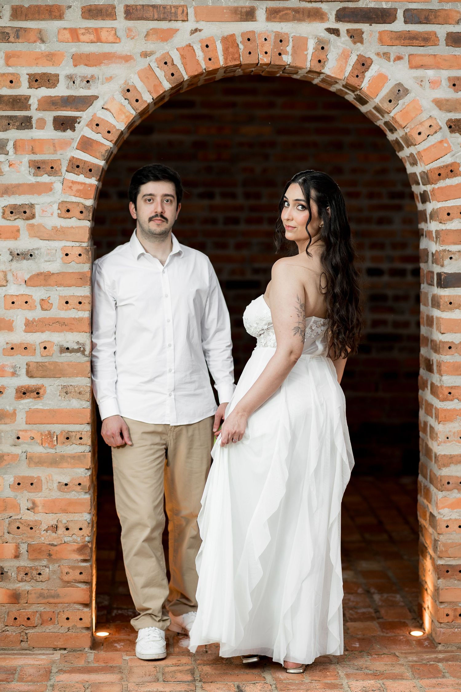 Couple posing in a rustic brick archway, man in white shirt and woman in elegant white bridal gown.