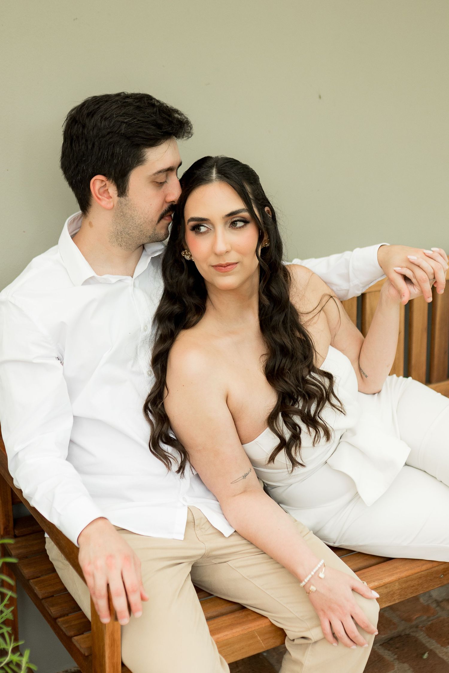 A couple in white and beige outfits sit closely on a wooden bench, the man kissing the woman's head tenderly.