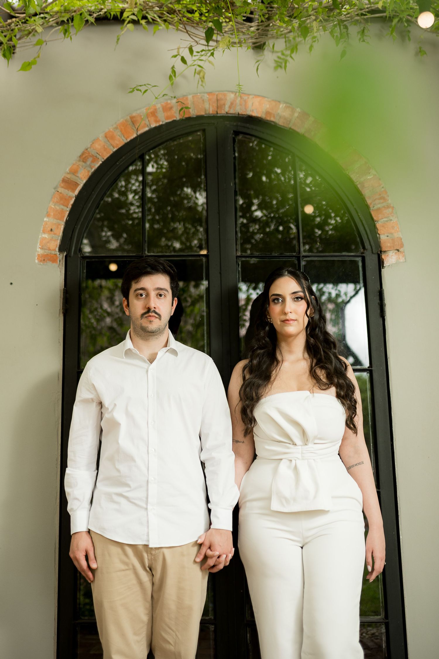 Couple in white outfits holding hands in front of an arched black door with brick accents and lush greenery overhead.