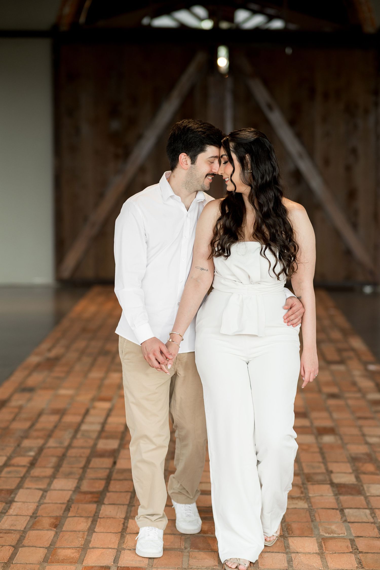 A couple in white and beige outfits share a romantic moment walking through a rustic barn venue.