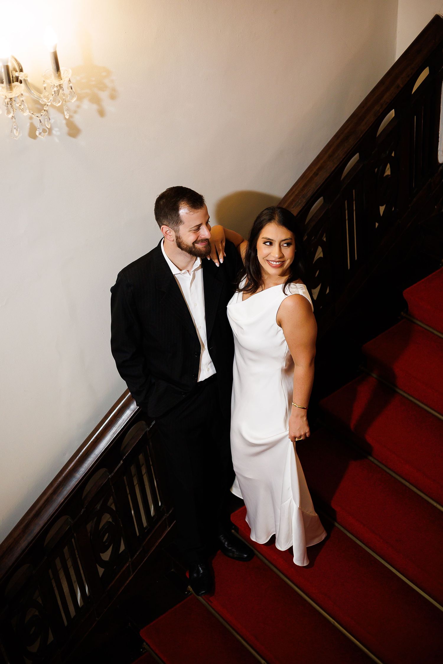 Elegant couple in formal attire posing on a grand staircase with red carpet and wooden railings.