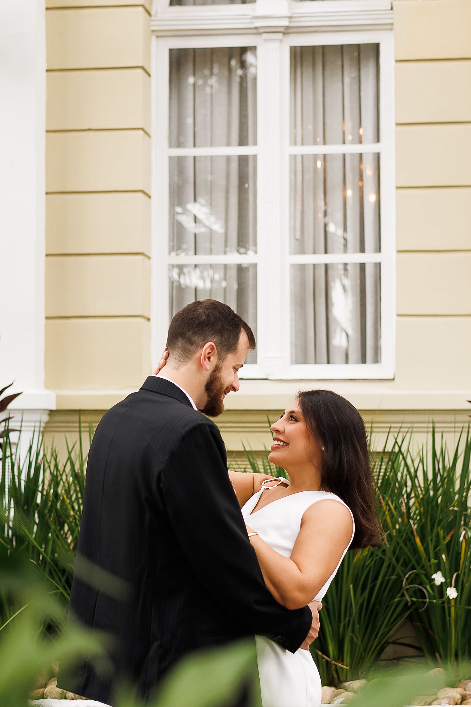 A bride and groom share a romantic moment outdoors, smiling at each other in front of a yellow building with white windows.