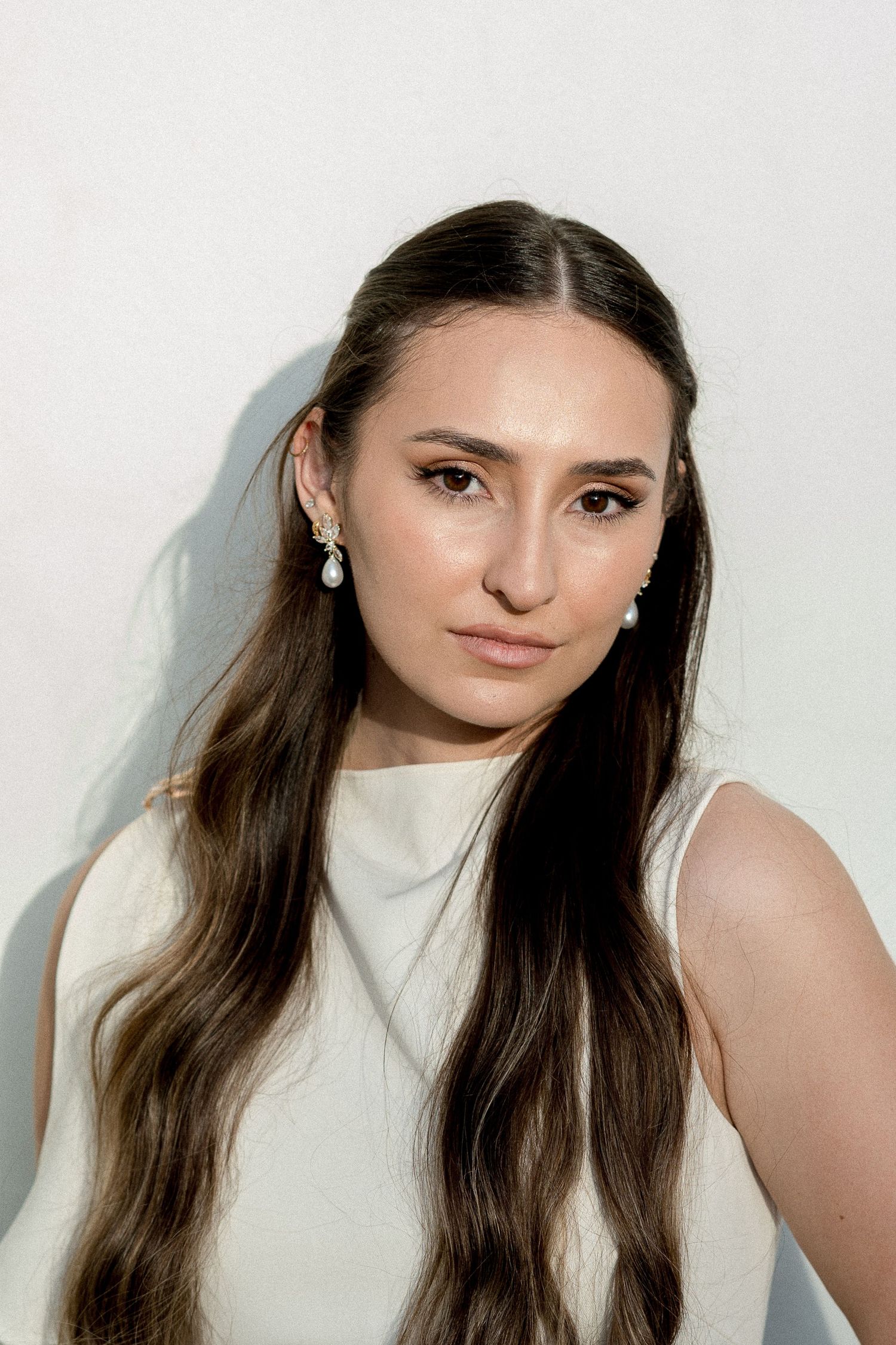 Young woman with long brown hair wearing pearl drop earrings and a white sleeveless top against a light background.