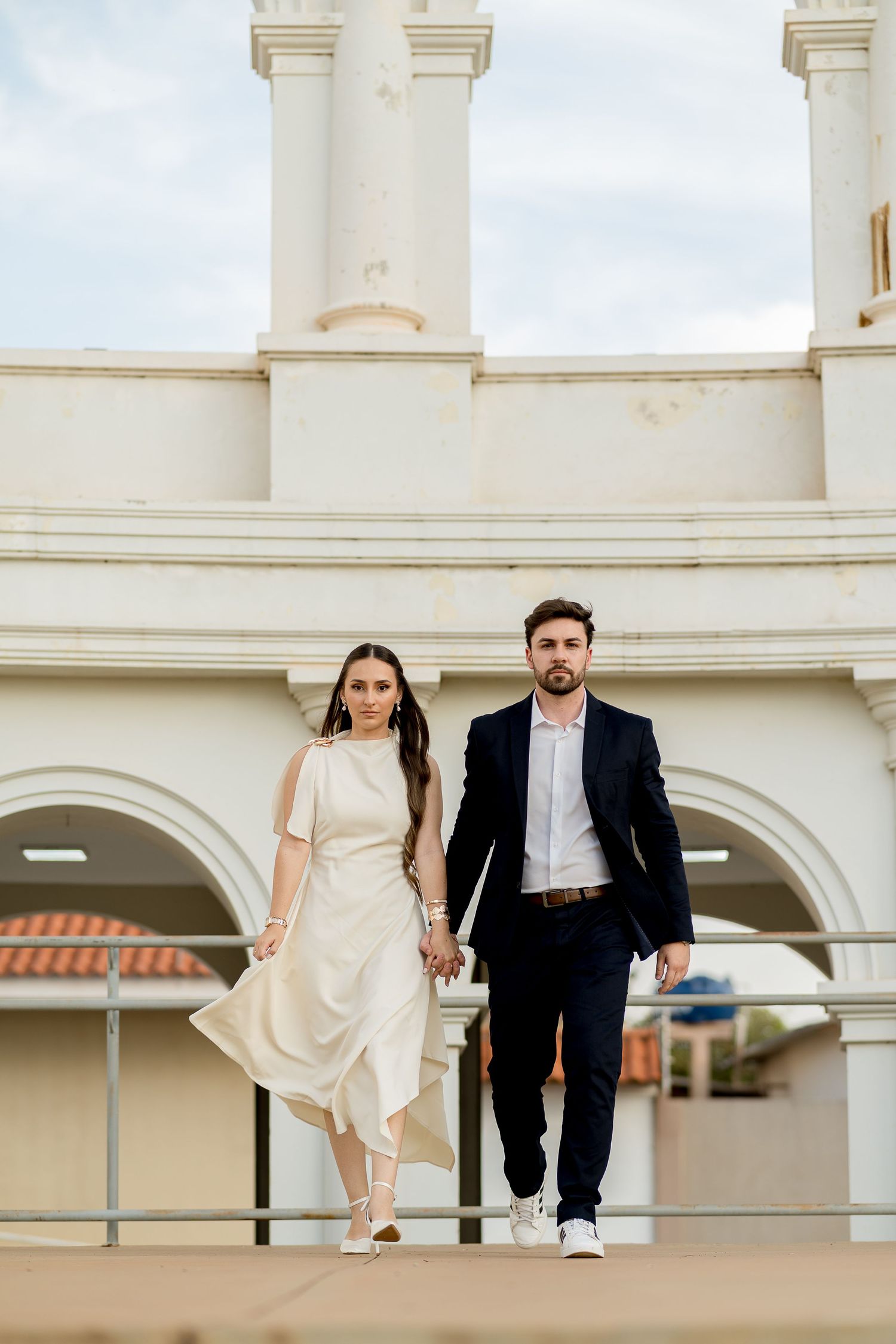 Elegant couple holding hands walking in front of white arched building, woman in white dress, man in navy suit.