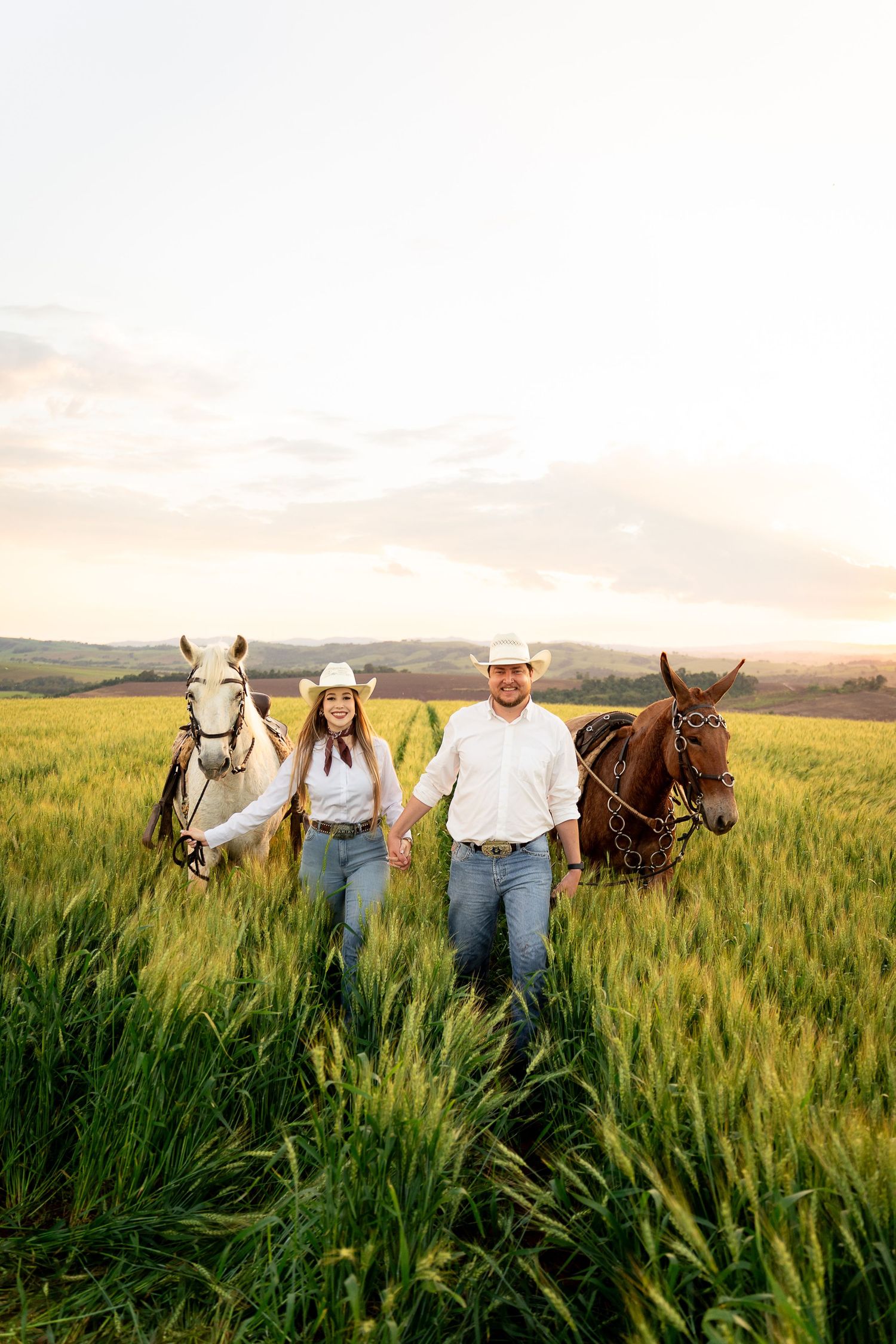 A couple in white shirts and cowboy hats hold hands walking through a green field with two horses at sunset.
