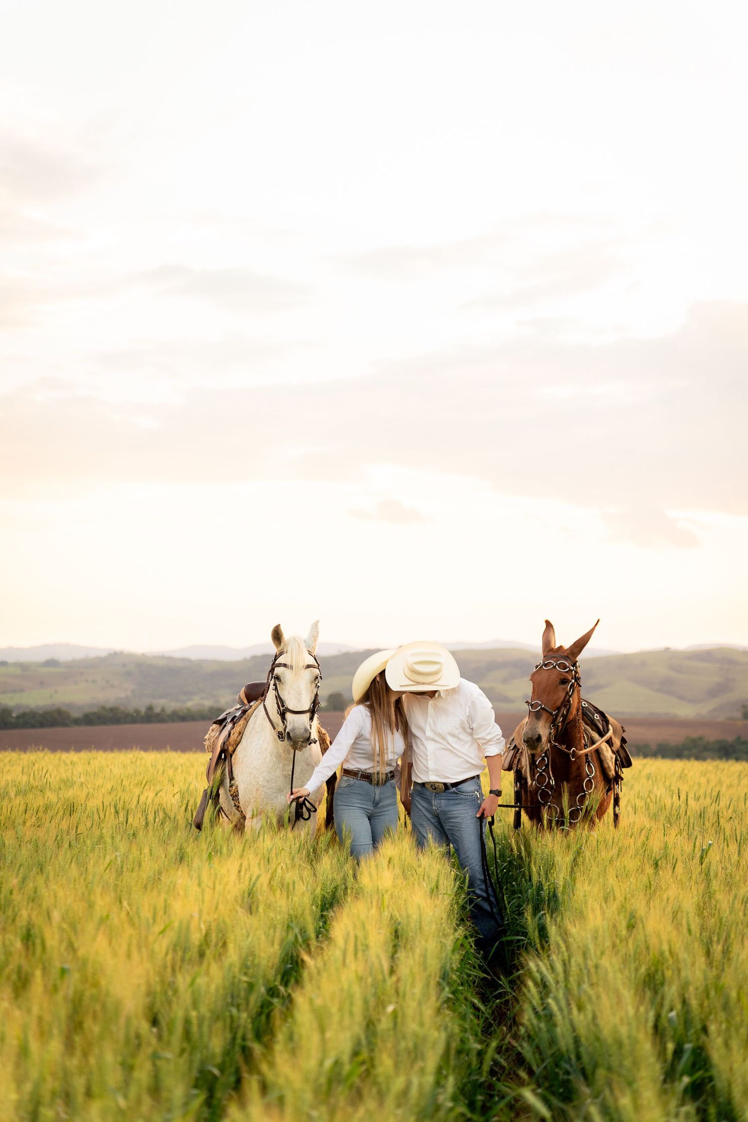 A cowgirl leads two horses, one white and one brown, through a lush green field at sunset.