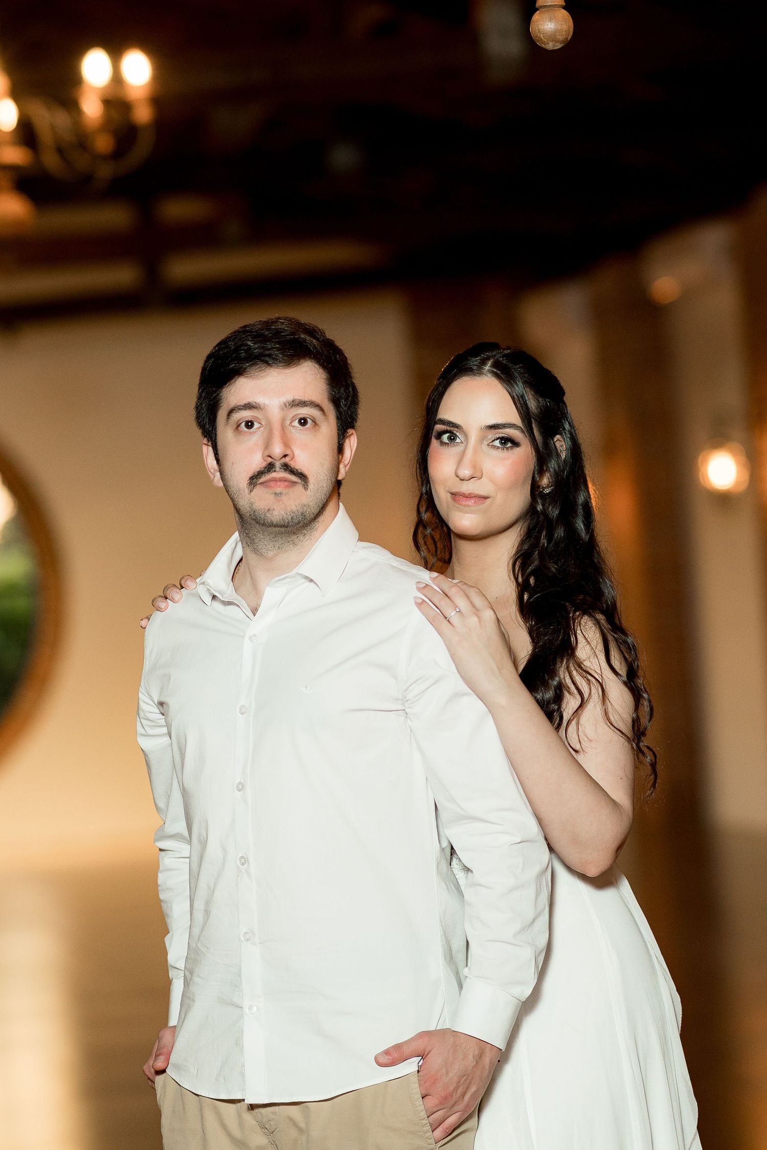 A couple in white outfits pose elegantly in a warmly lit venue, the woman placing her hand on the mans shoulder.