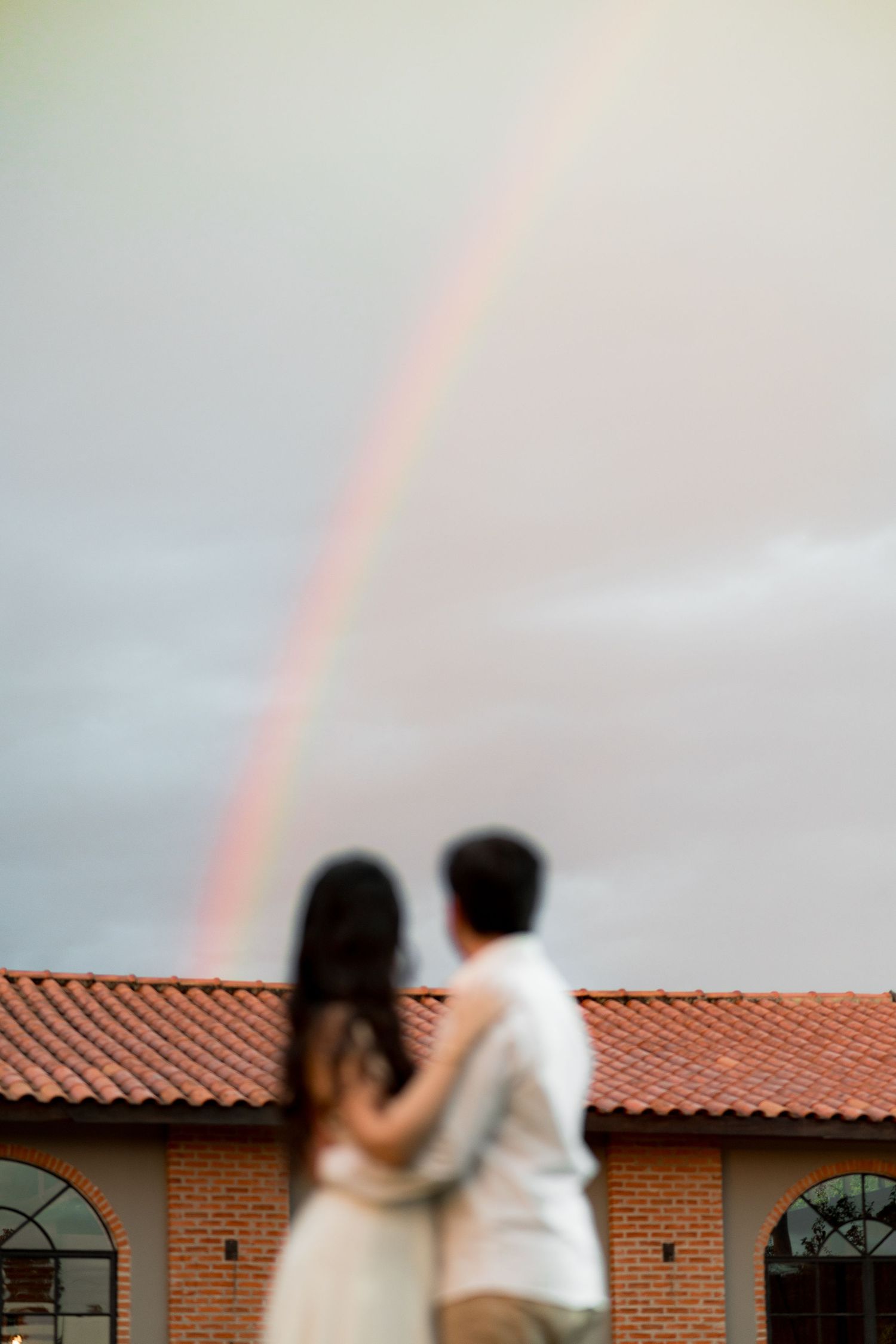 Couple embracing outdoors with a vibrant double rainbow arching over a red-tiled roof building in the background.