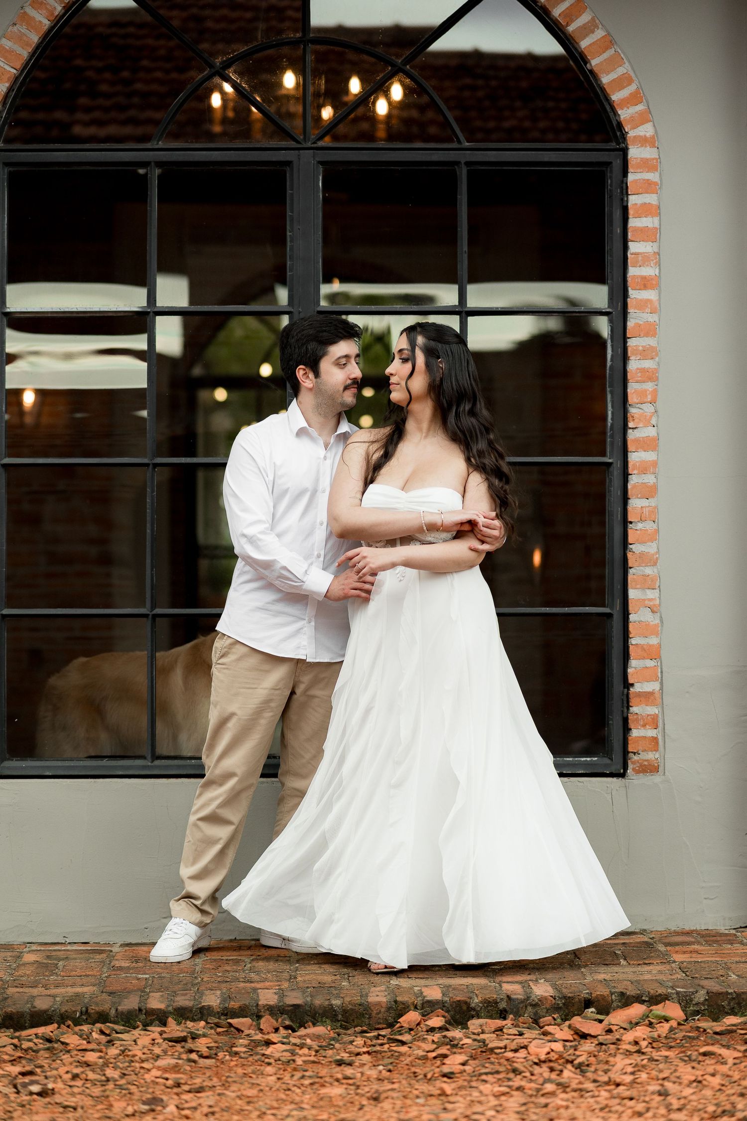 Couple in wedding attire embracing outdoors near arched window, bride in white gown, groom in white shirt and khakis.