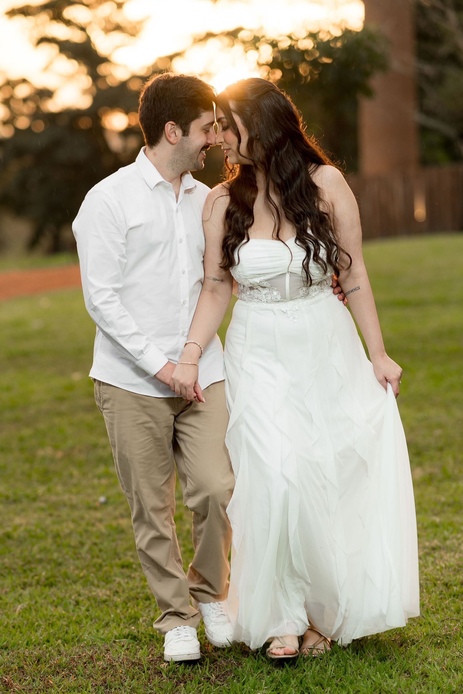 Couple in wedding attire sharing a romantic moment at sunset on a green lawn, woman in white gown, man in white shirt.