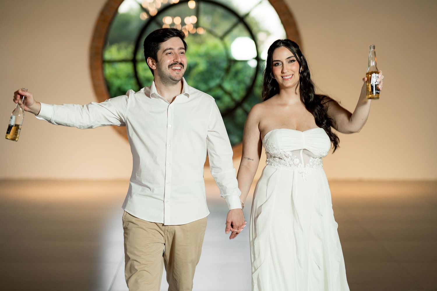 Smiling couple in wedding attire holding beer bottles in elegant venue with round window backdrop.