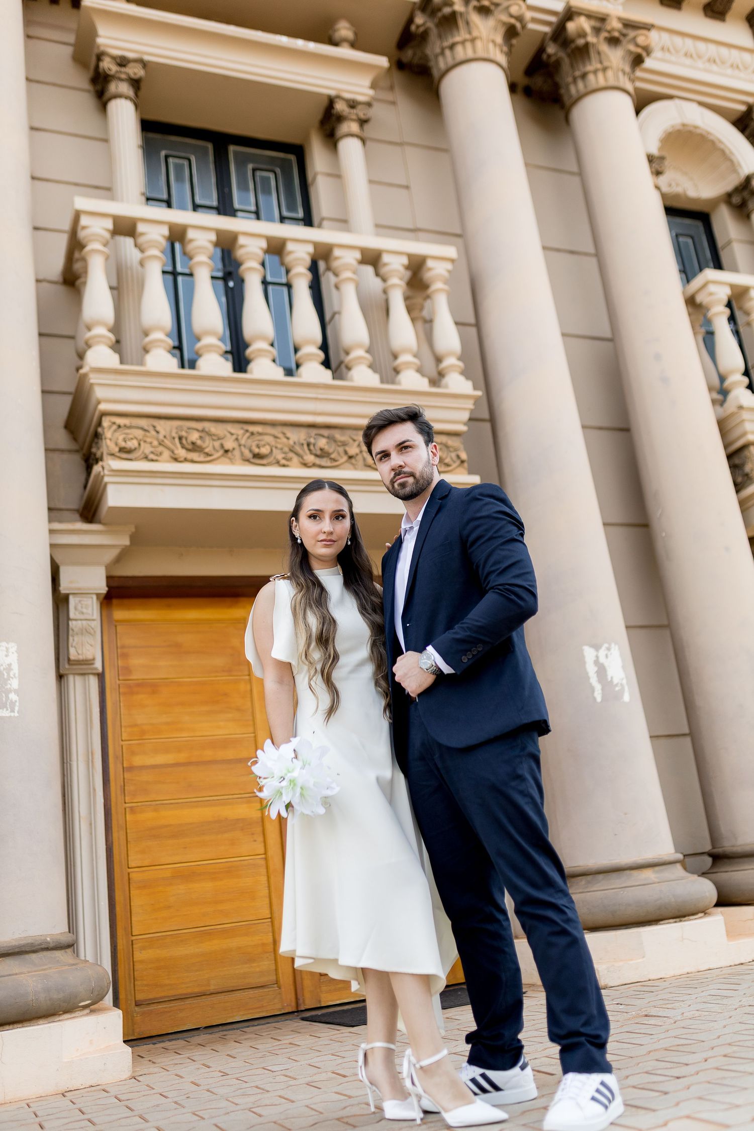 Elegant couple in wedding attire posing before a classical building with ornate columns and balustrade.