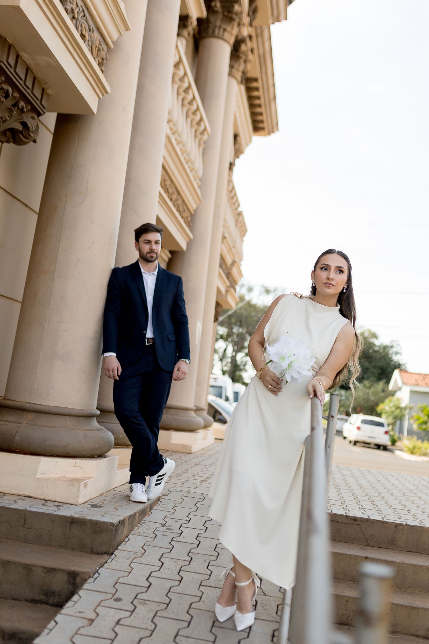 Elegant bride in white dress holding bouquet poses on steps with groom in navy suit beside classical columns.