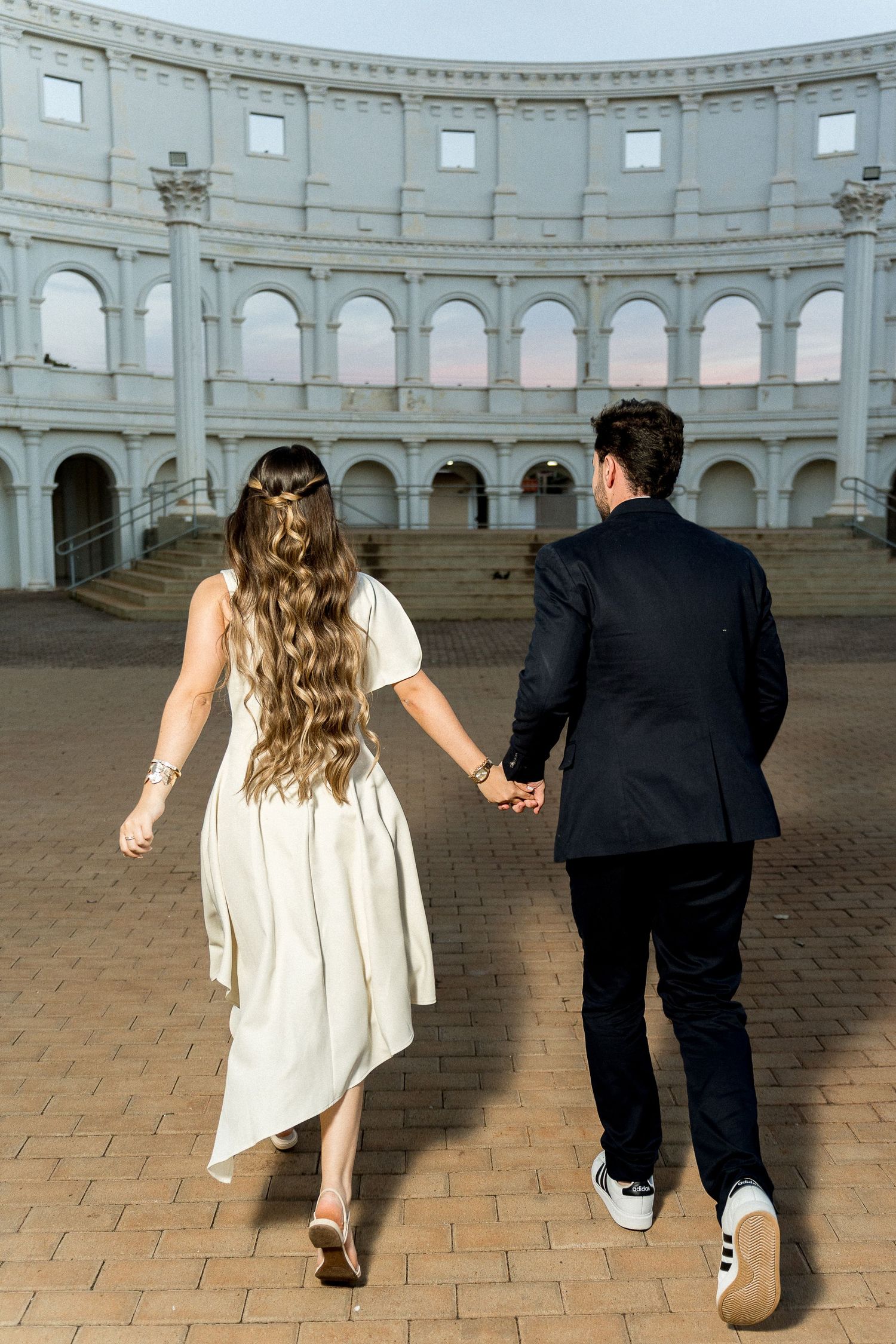 A couple in wedding attire holds hands walking toward a grand Roman-style amphitheater at dusk.