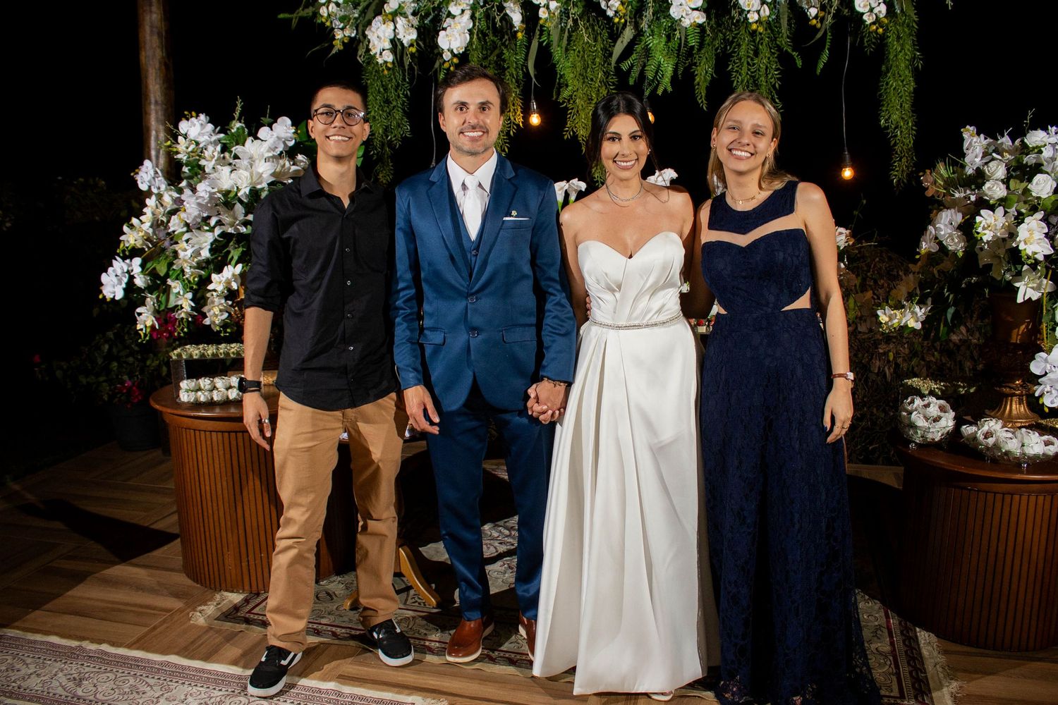 Wedding party of four posing outdoors at night with floral arch backdrop, bride in white gown and groom in blue suit.