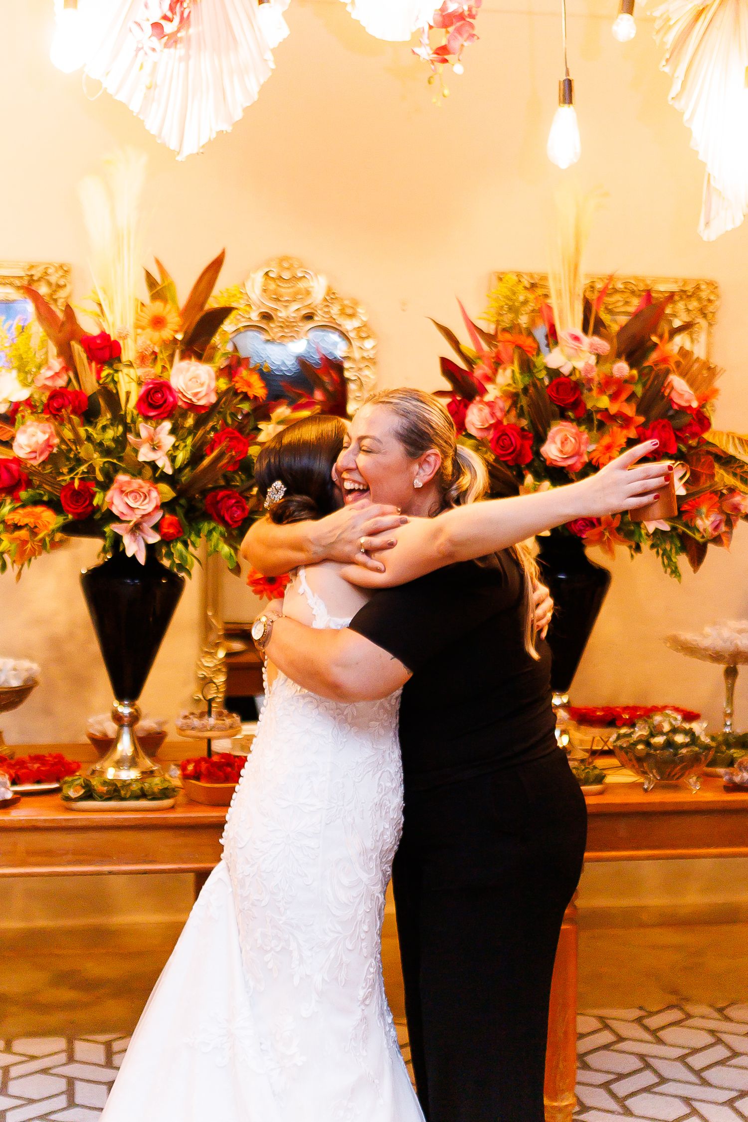 Bride in white lace gown hugging woman in black dress at wedding reception with floral arrangements.