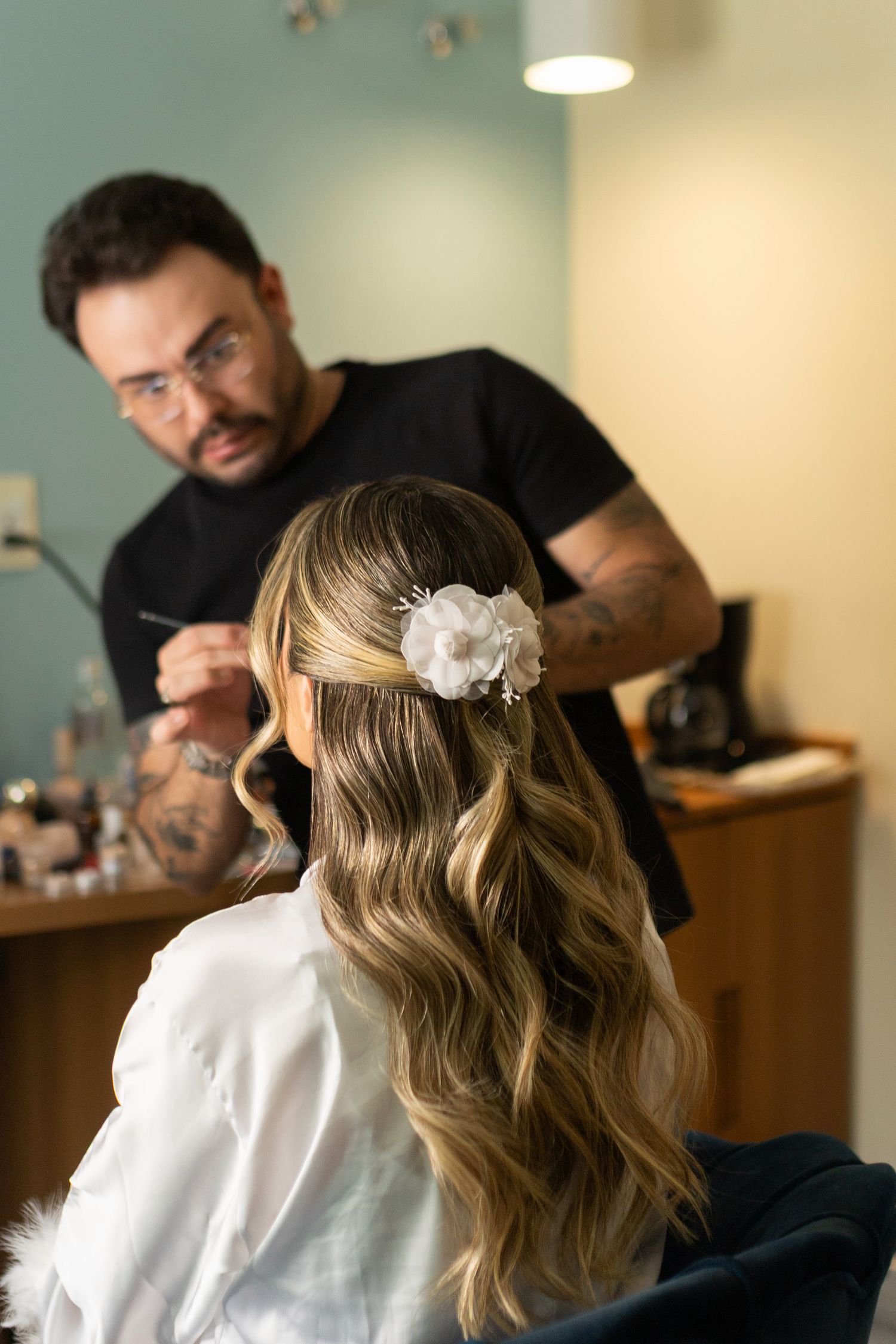 A hairstylist with tattoos applies finishing touches to a bride's elegant half-up wavy hairstyle adorned with a white flower.