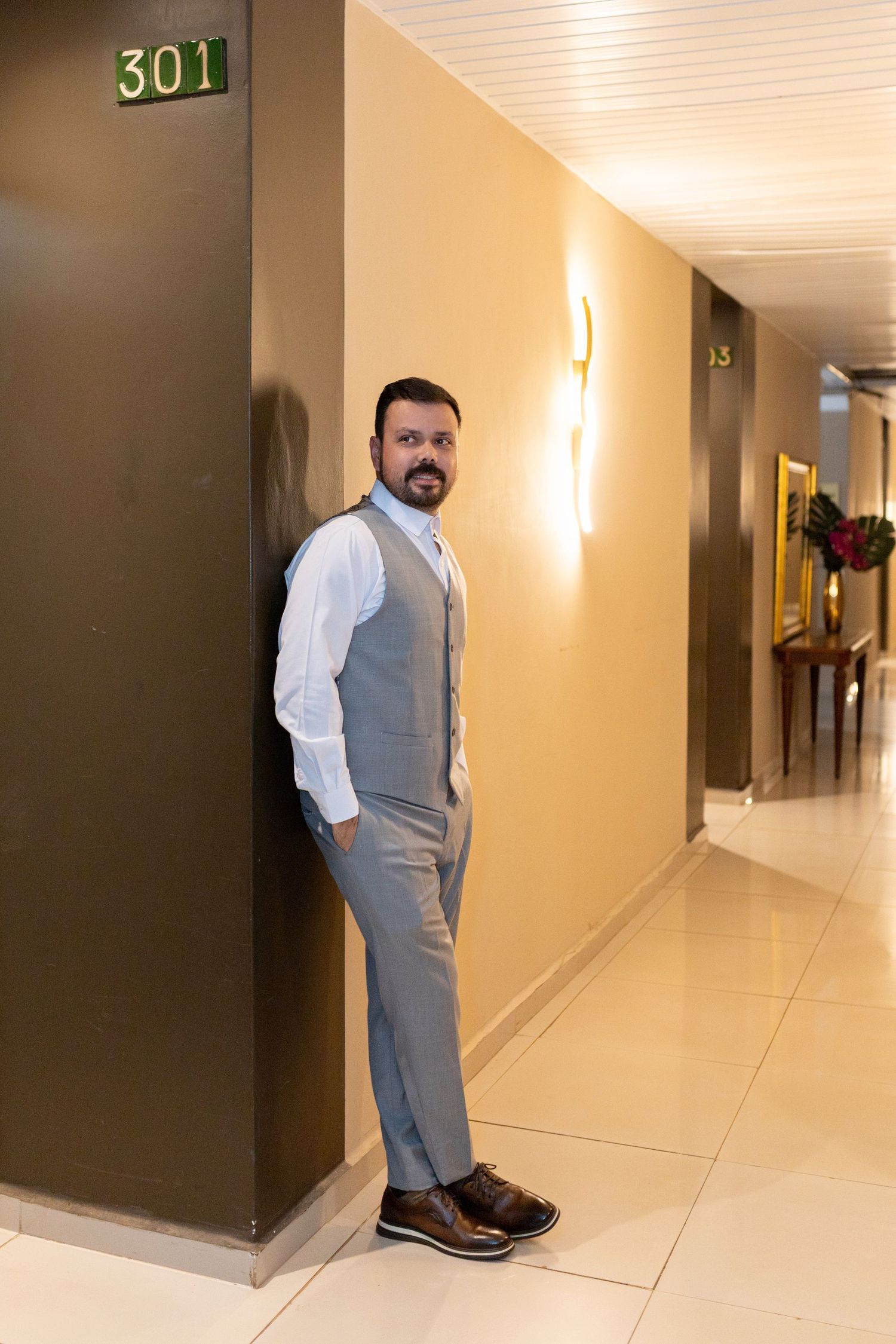 Man in gray vest and trousers leaning against hotel corridor wall near room 301 sign.