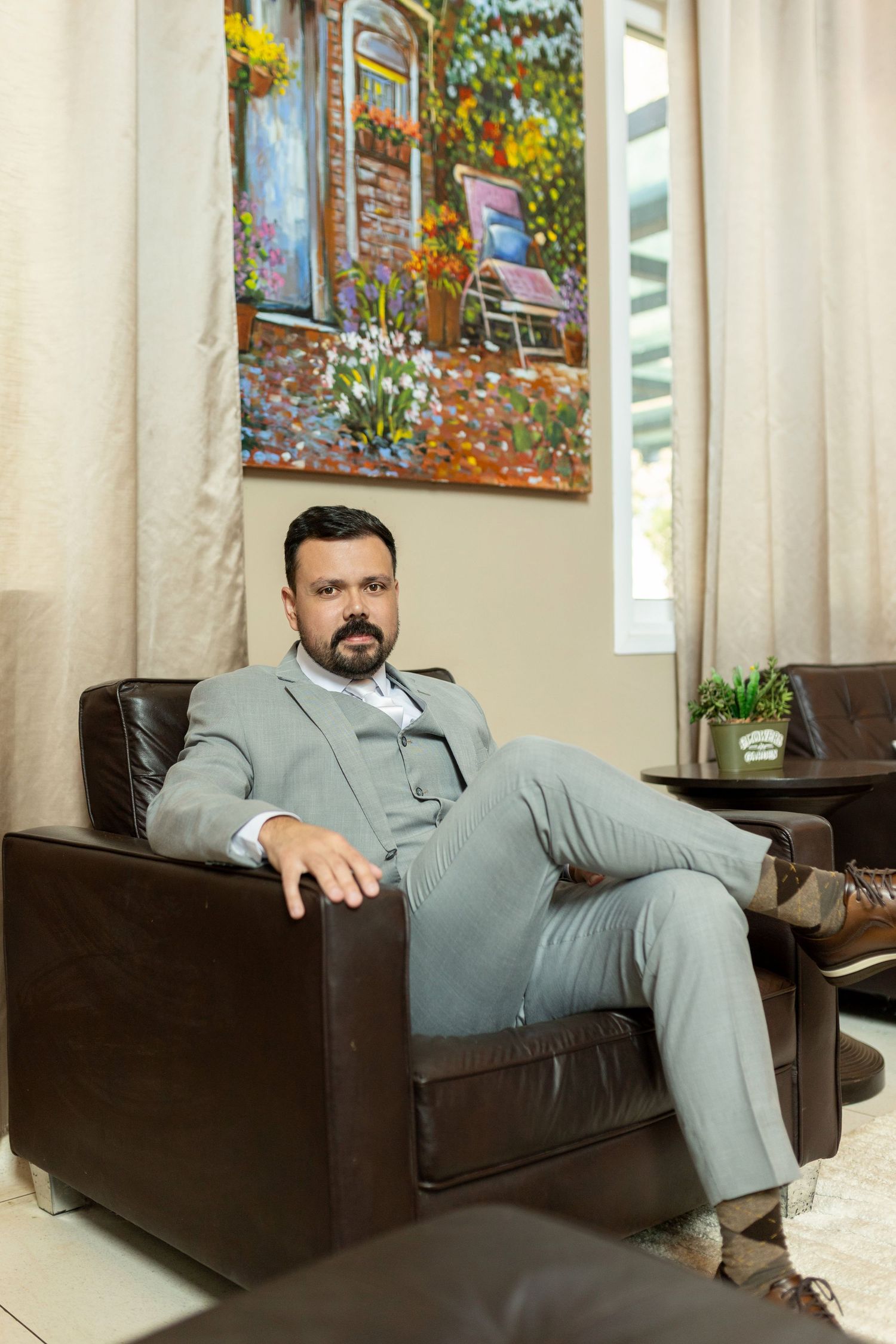 Professional man in gray suit sitting relaxed in brown leather armchair with colorful painting on wall behind him.
