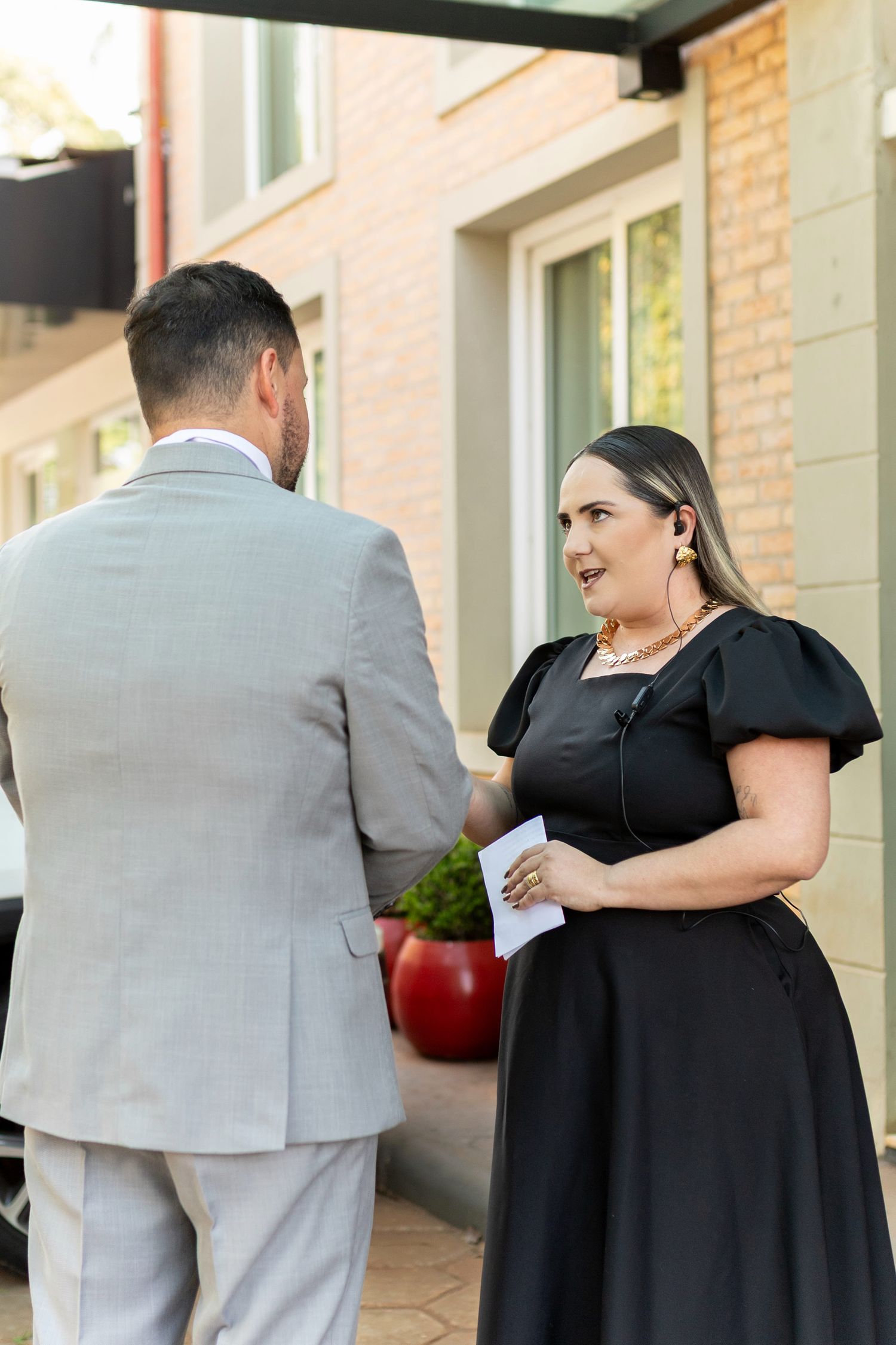 A woman in a black dress with puff sleeves speaks with a man in a gray suit outside a brick building.