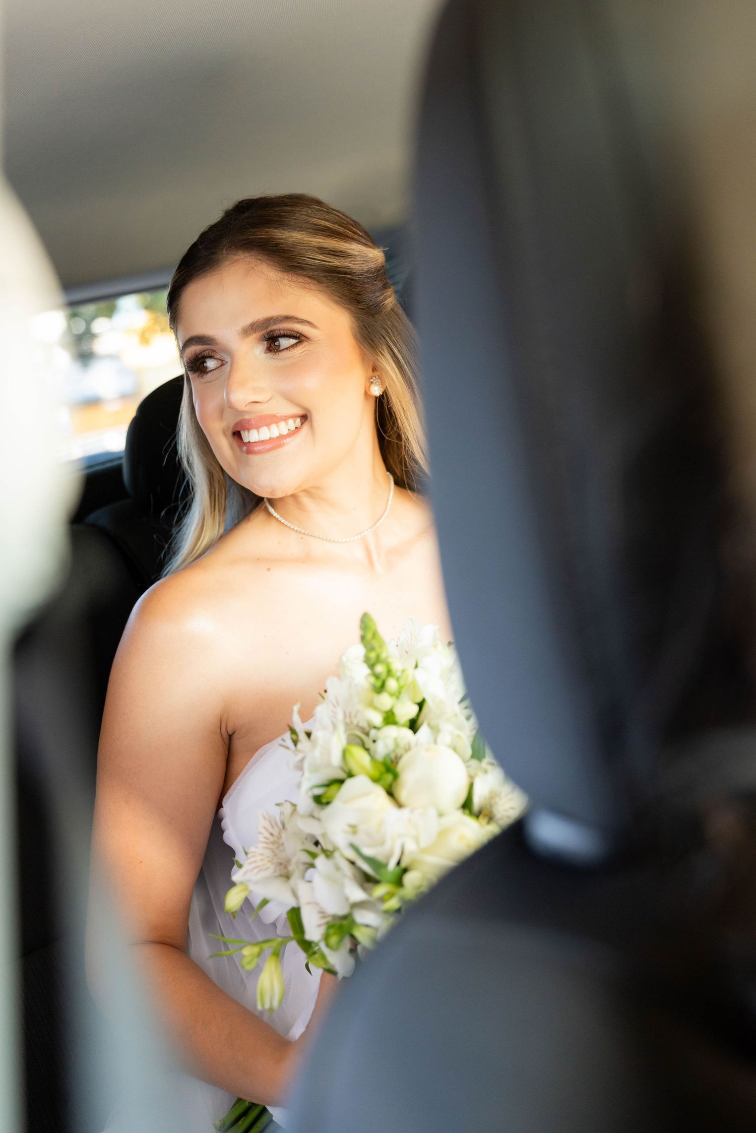 Smiling bride in strapless gown holding white floral bouquet while seated in a car on her wedding day.