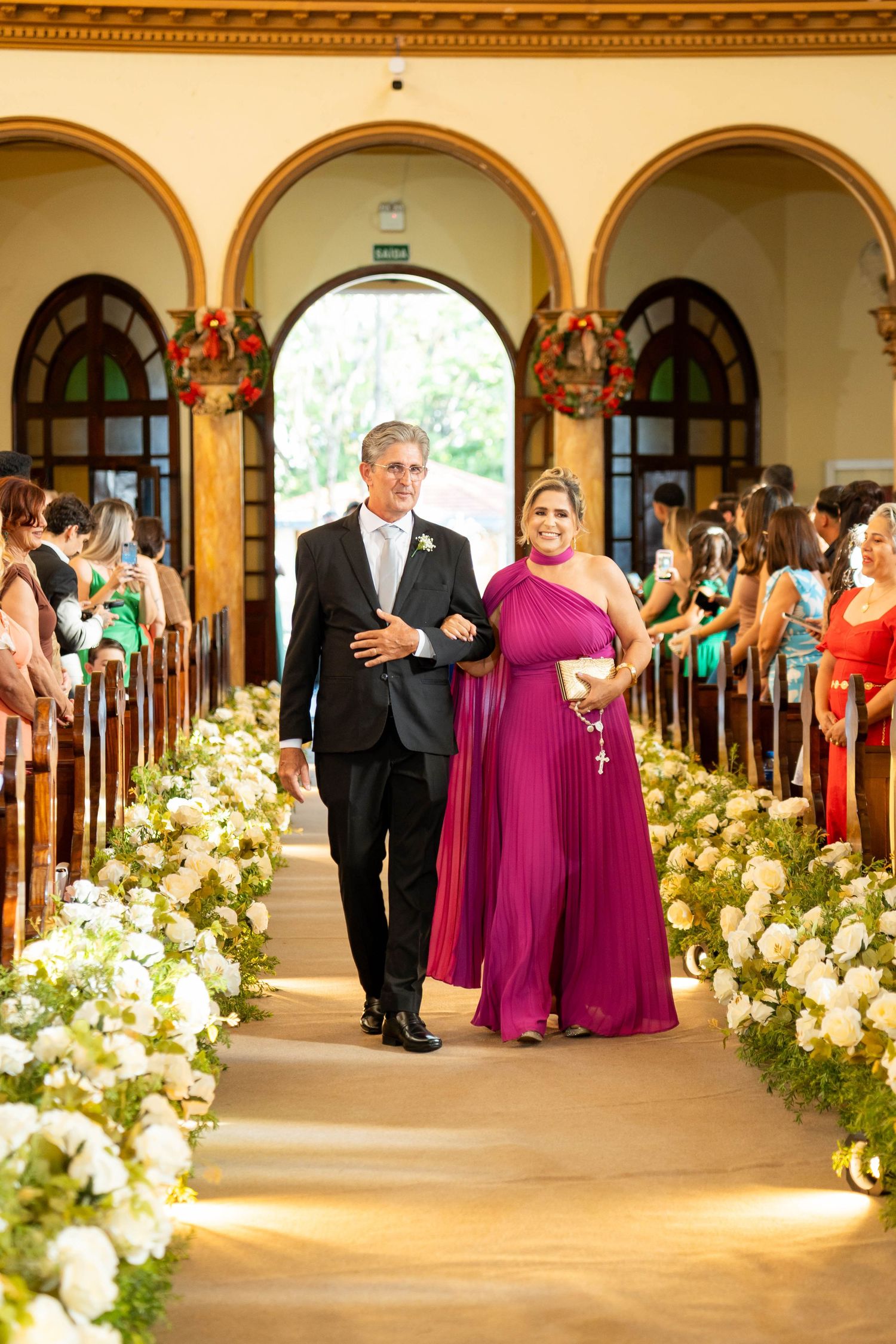 Elegant couple walks down wedding aisle lined with white flowers, woman in purple gown holding rosary.