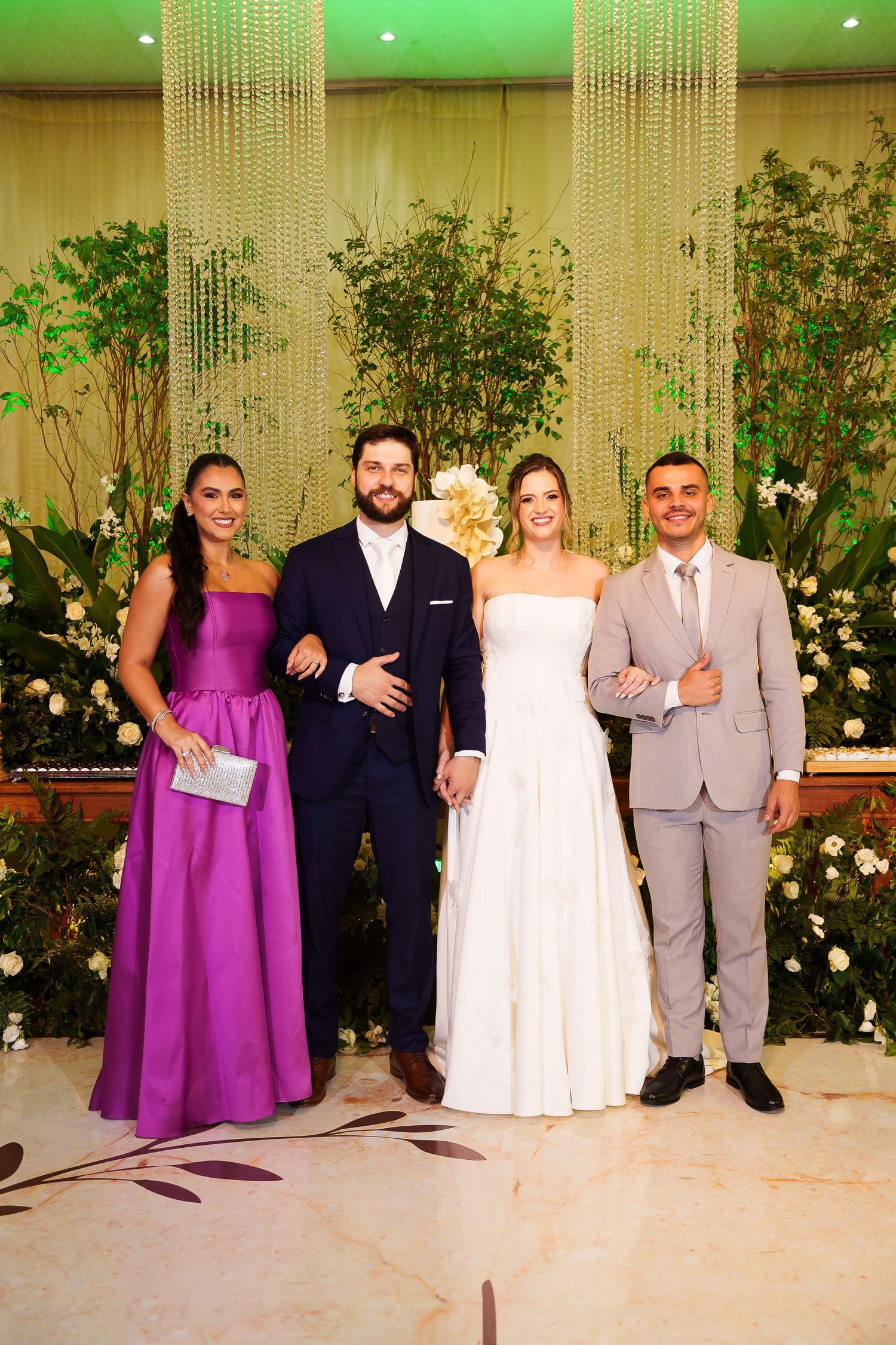 Four elegantly dressed wedding guests pose together in front of a floral backdrop at a formal wedding reception.