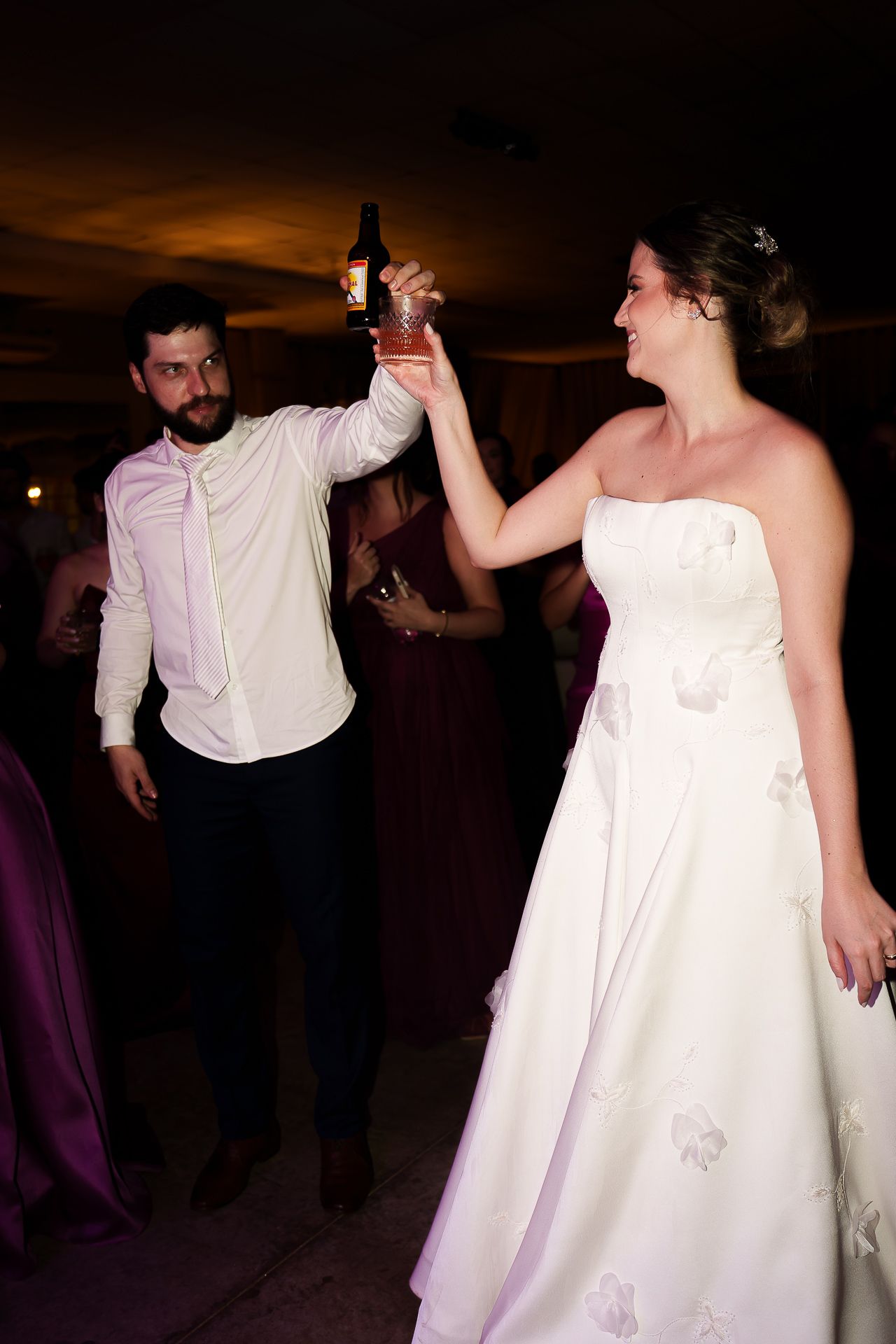 Bride in white gown and groom in white shirt dancing together, raising a bottle at a wedding reception.